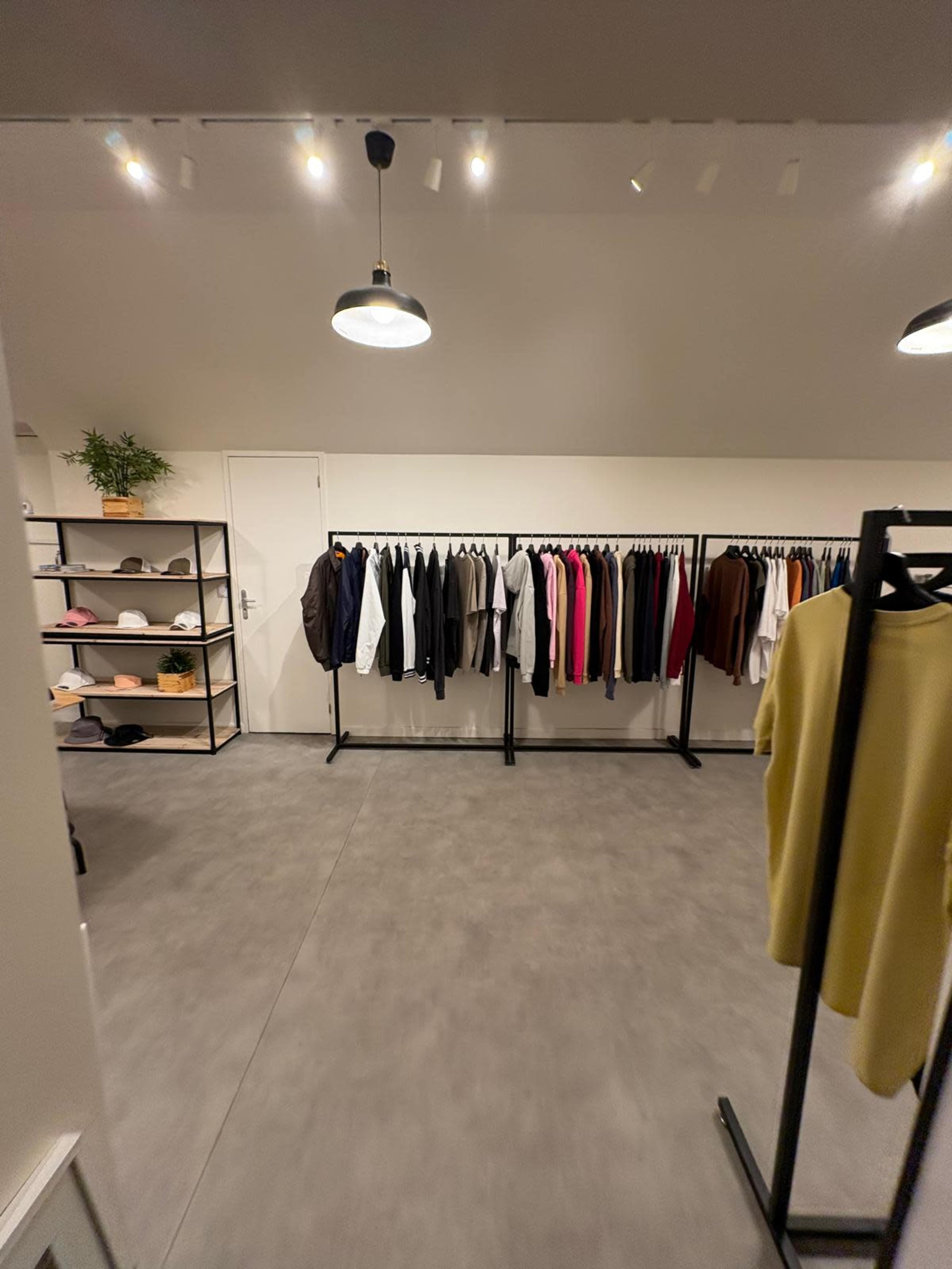 The image shows a clothing display area with racks of assorted garments organized neatly along the walls and a shelf with accessories in the corner.