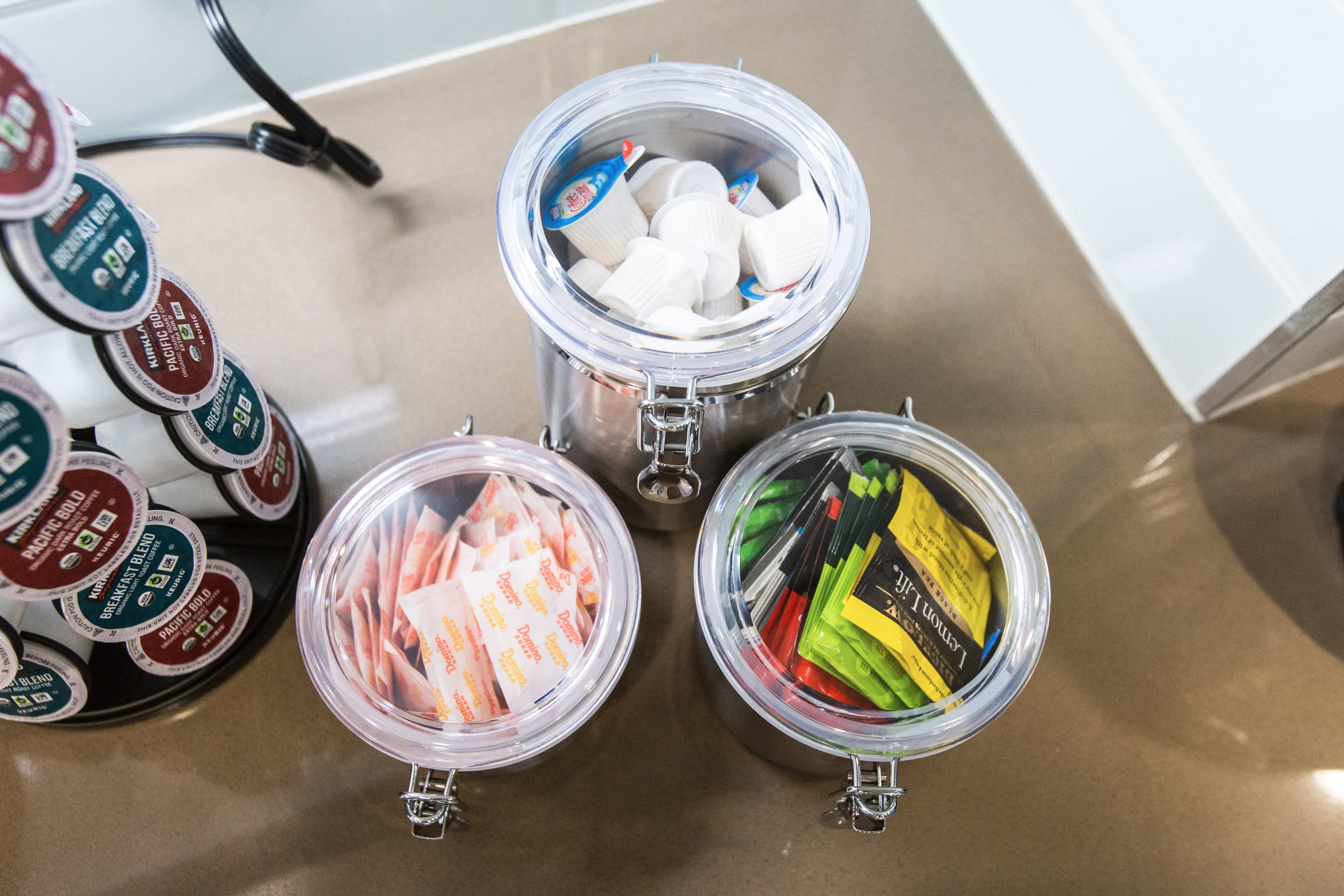 Three clear jars filled with various coffee condiments, including sugar packets, creamers, and stirrers, arranged on a countertop next to a coffee pod holder.