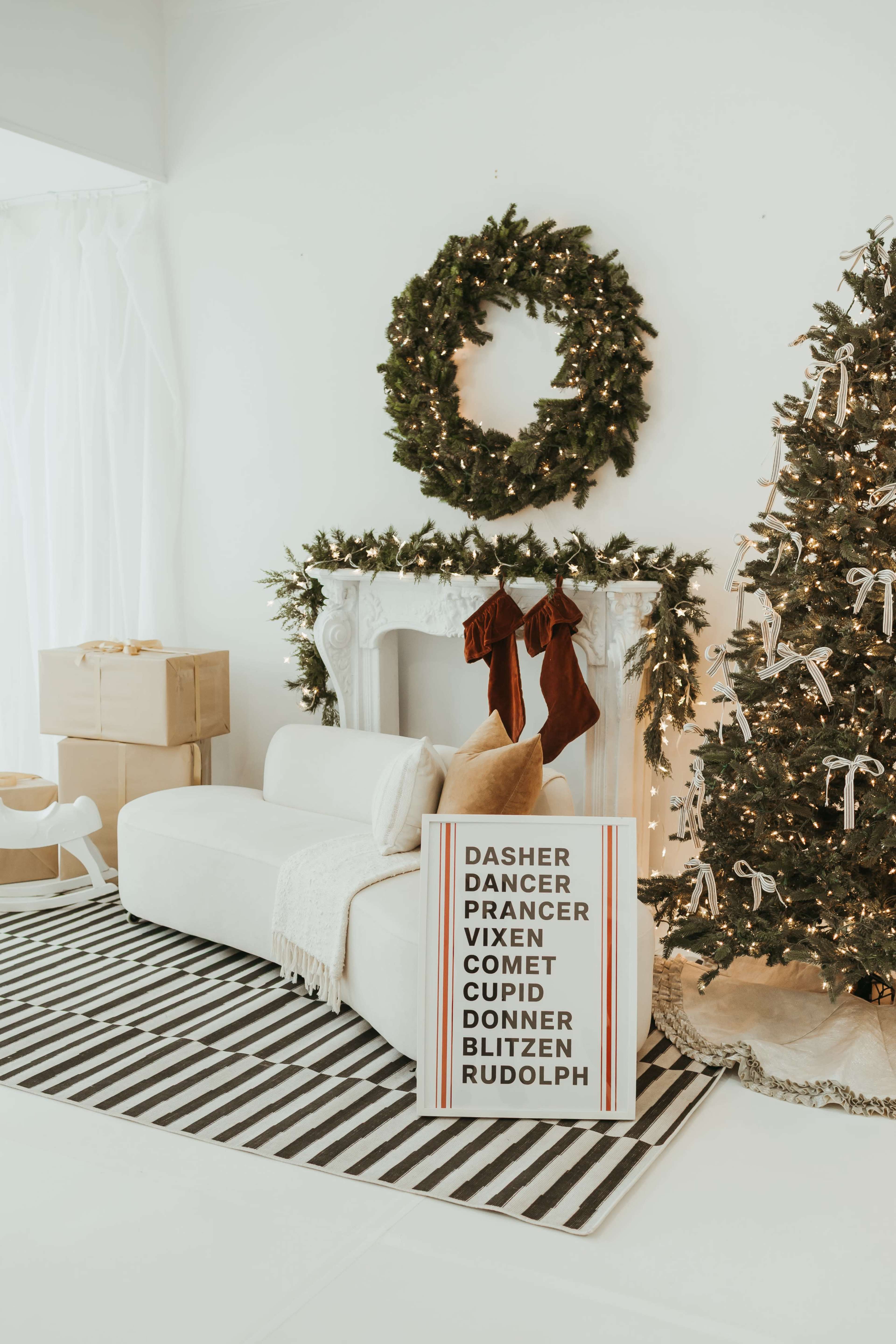 The image shows a decorated living room featuring a Christmas tree, stockings hanging from a white mantel, and a sign listing the names of Santa's reindeer.