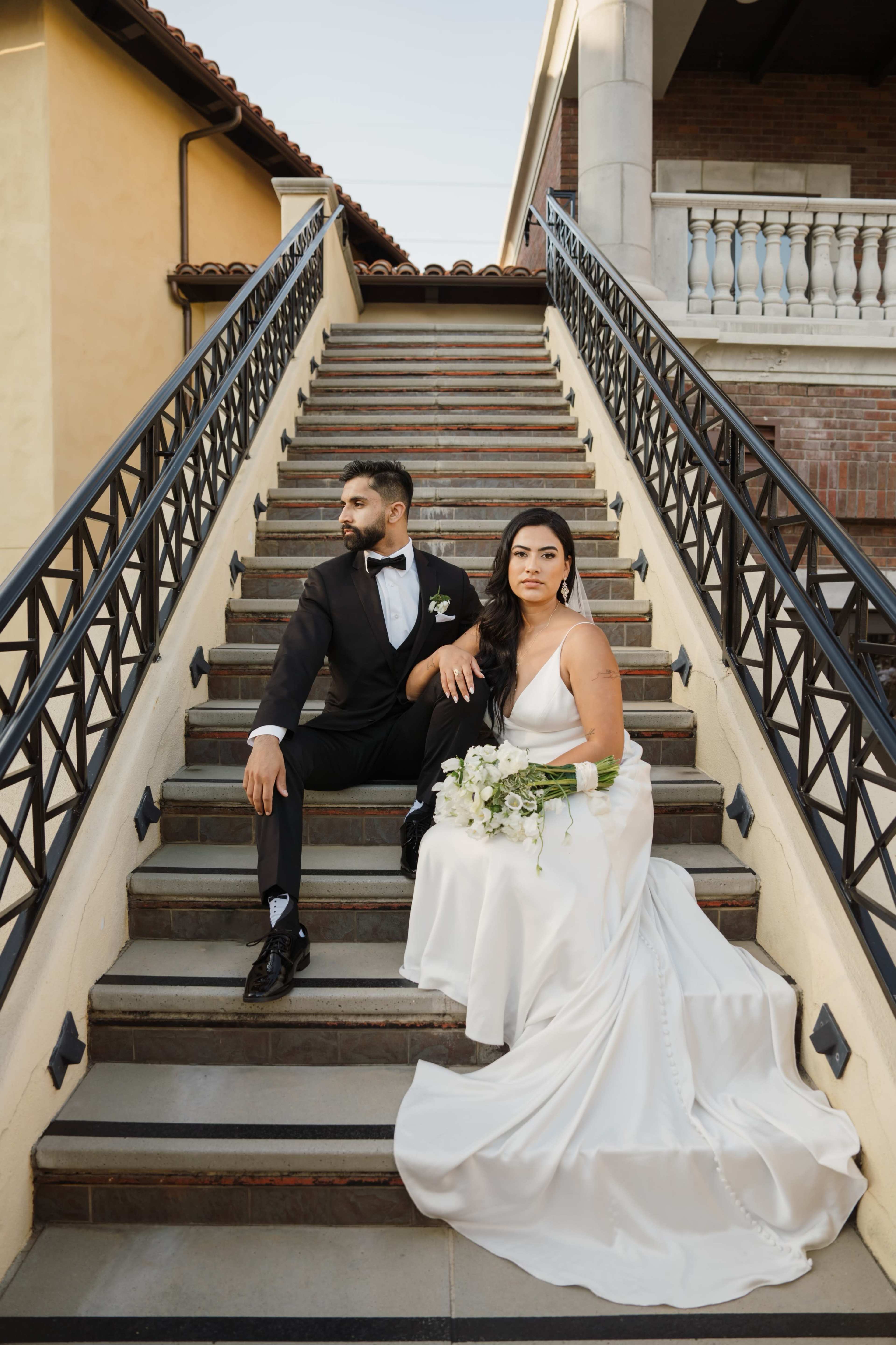 A bride and groom sit on a staircase, with the bride in a white gown and the groom in a black tuxedo, both looking away from each other.