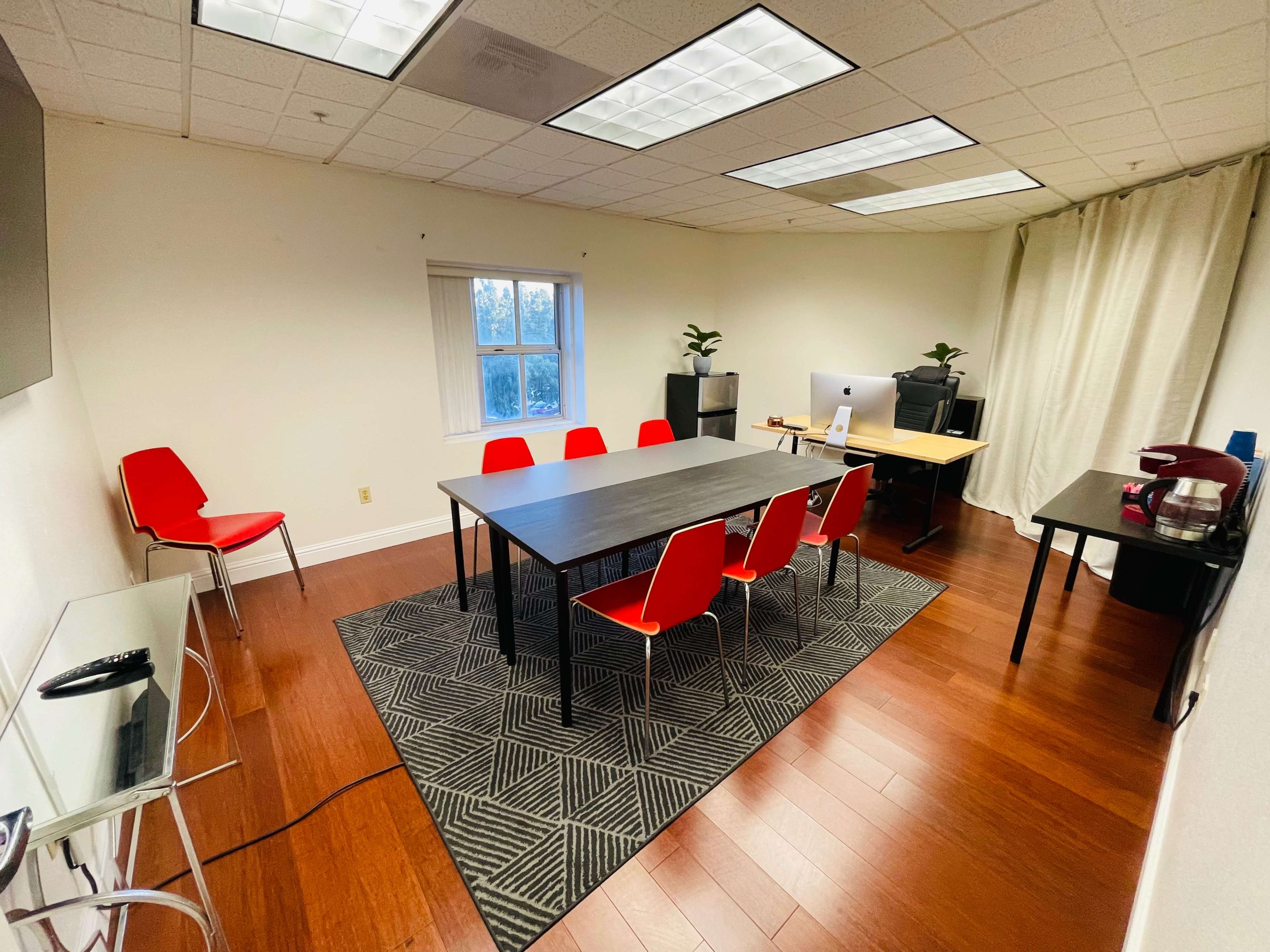 The image shows a meeting room with a large table surrounded by six red chairs, a window, a desk with a computer, and a coffee machine.