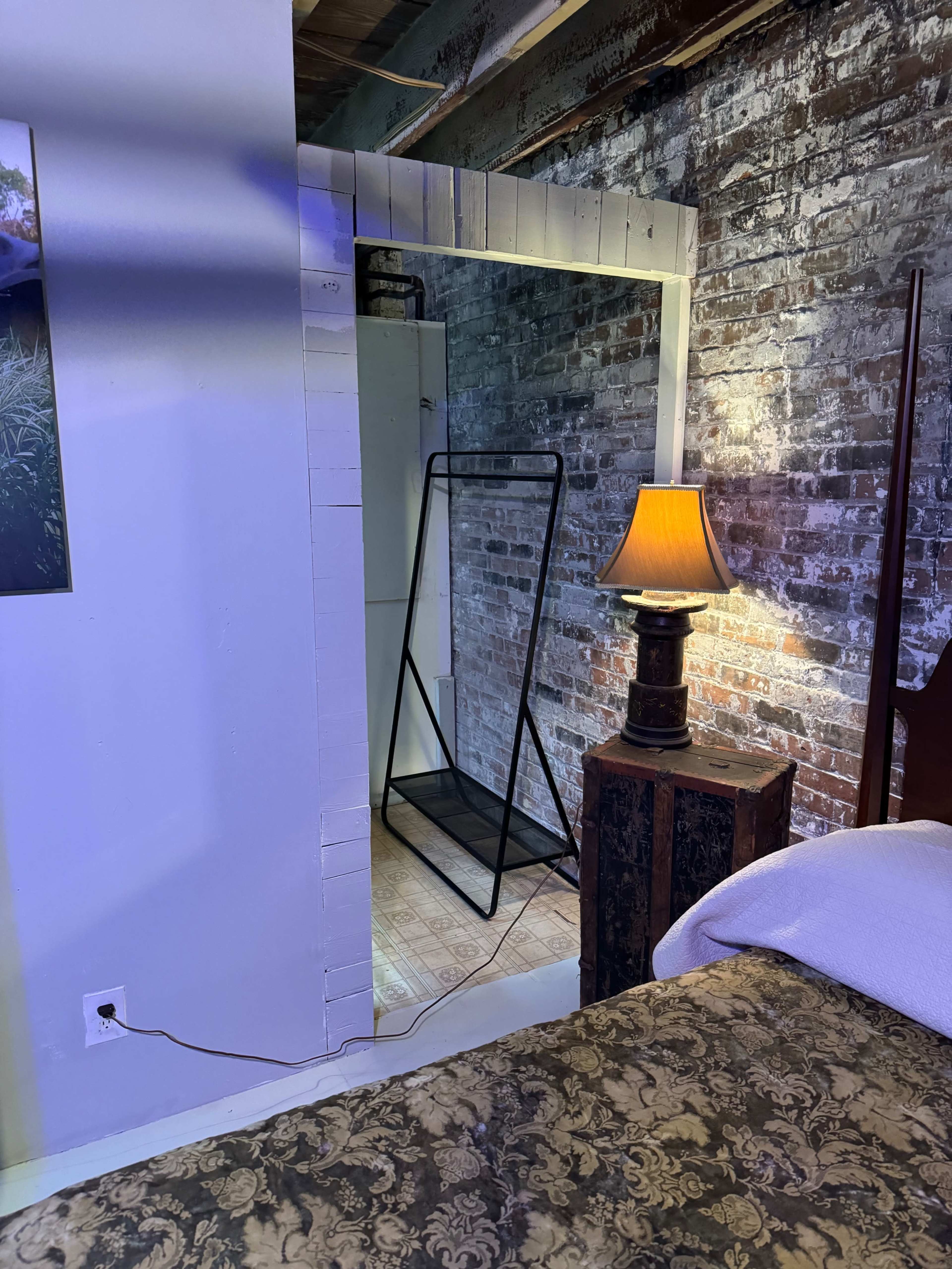 A small room features a bed with a patterned blanket, a wooden nightstand with a lamp, and a black clothing rack in an open space framed by exposed brick walls.