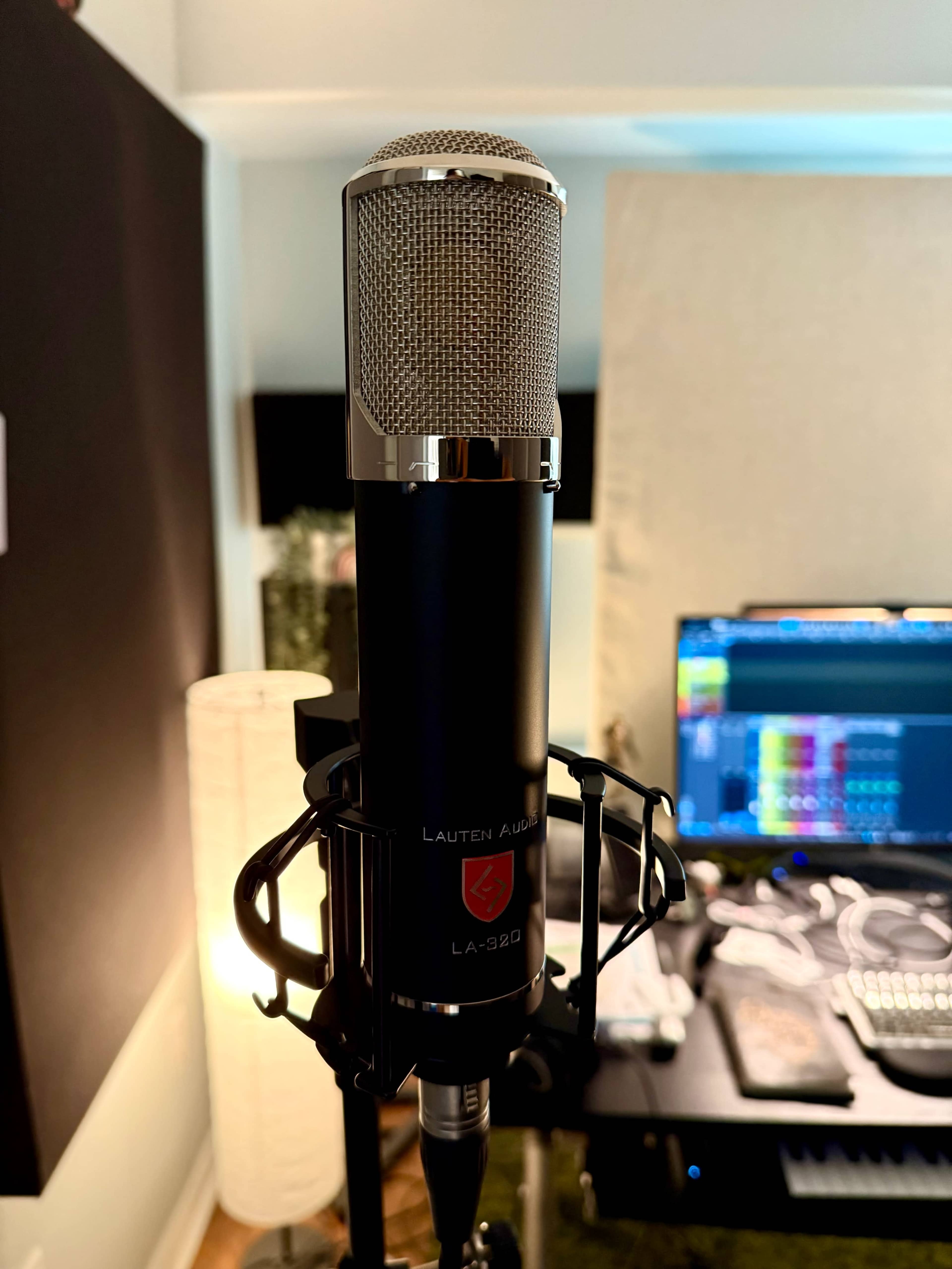 A Lauten Audio LA-920 microphone is mounted on a stand in a home recording studio, with a computer screen displaying audio software in the background.