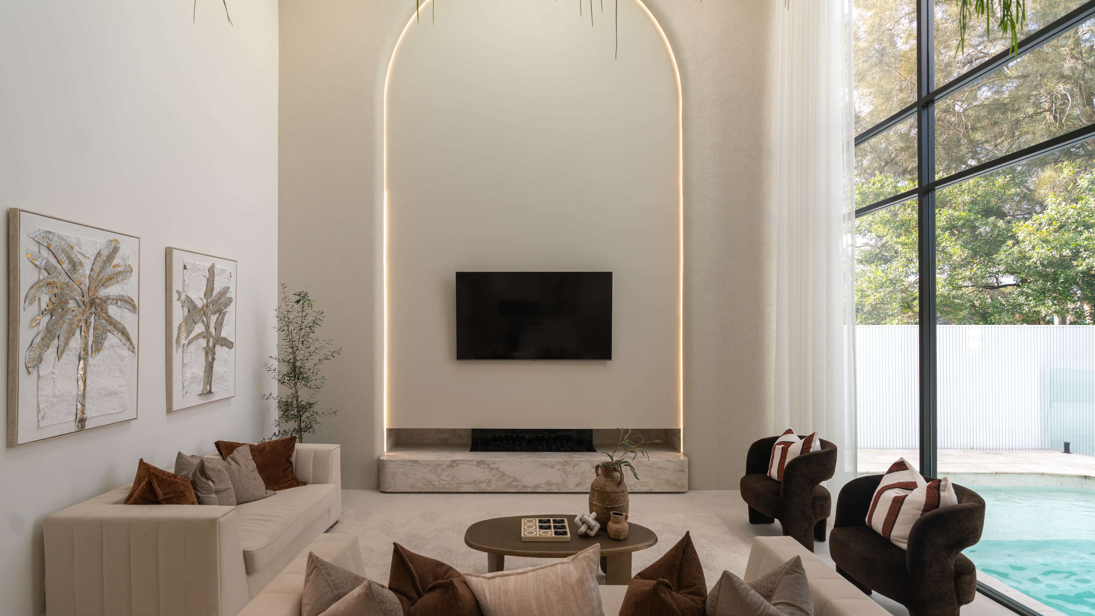 The image features a modern living room with a high ceiling, a minimalist fireplace below a wall-mounted TV, and large windows that overlook a pool area.