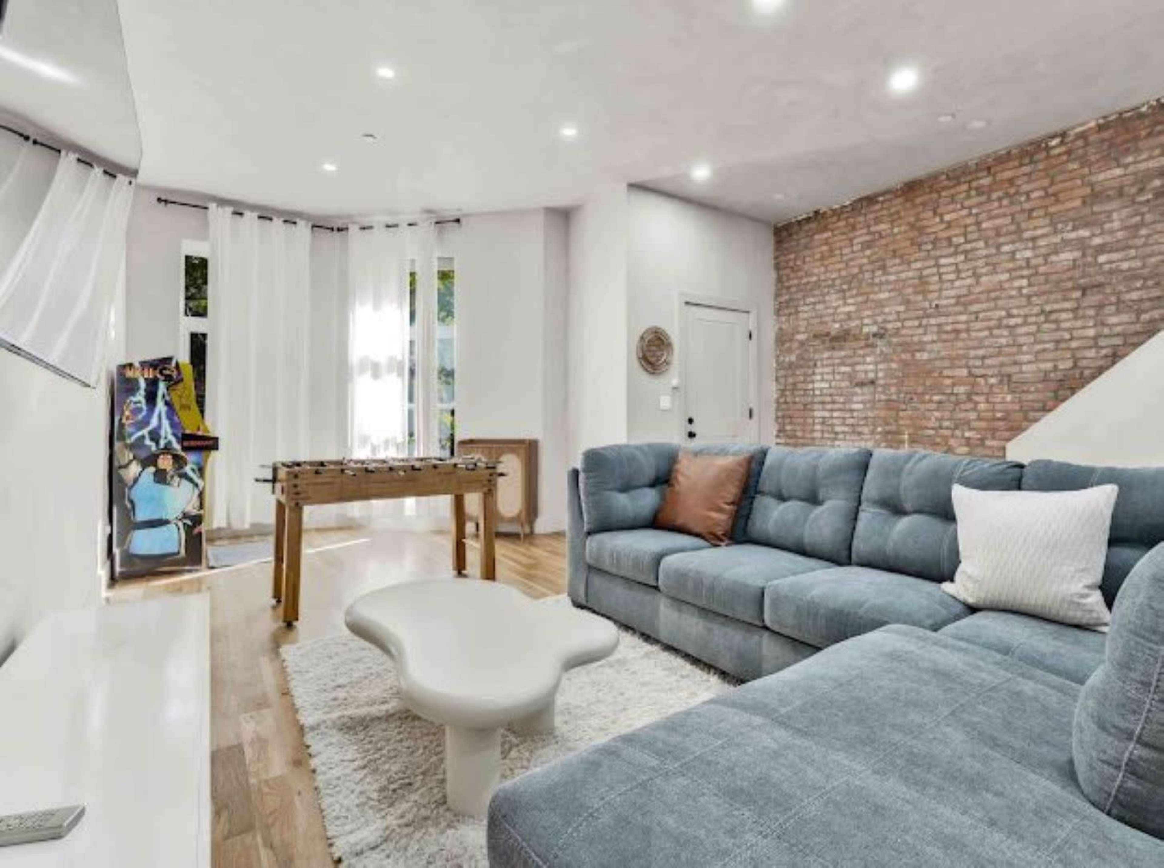 The image shows a modern living room with a blue sectional sofa, a round coffee table, and a foosball table, featuring a brick accent wall and large windows with sheer white curtains.