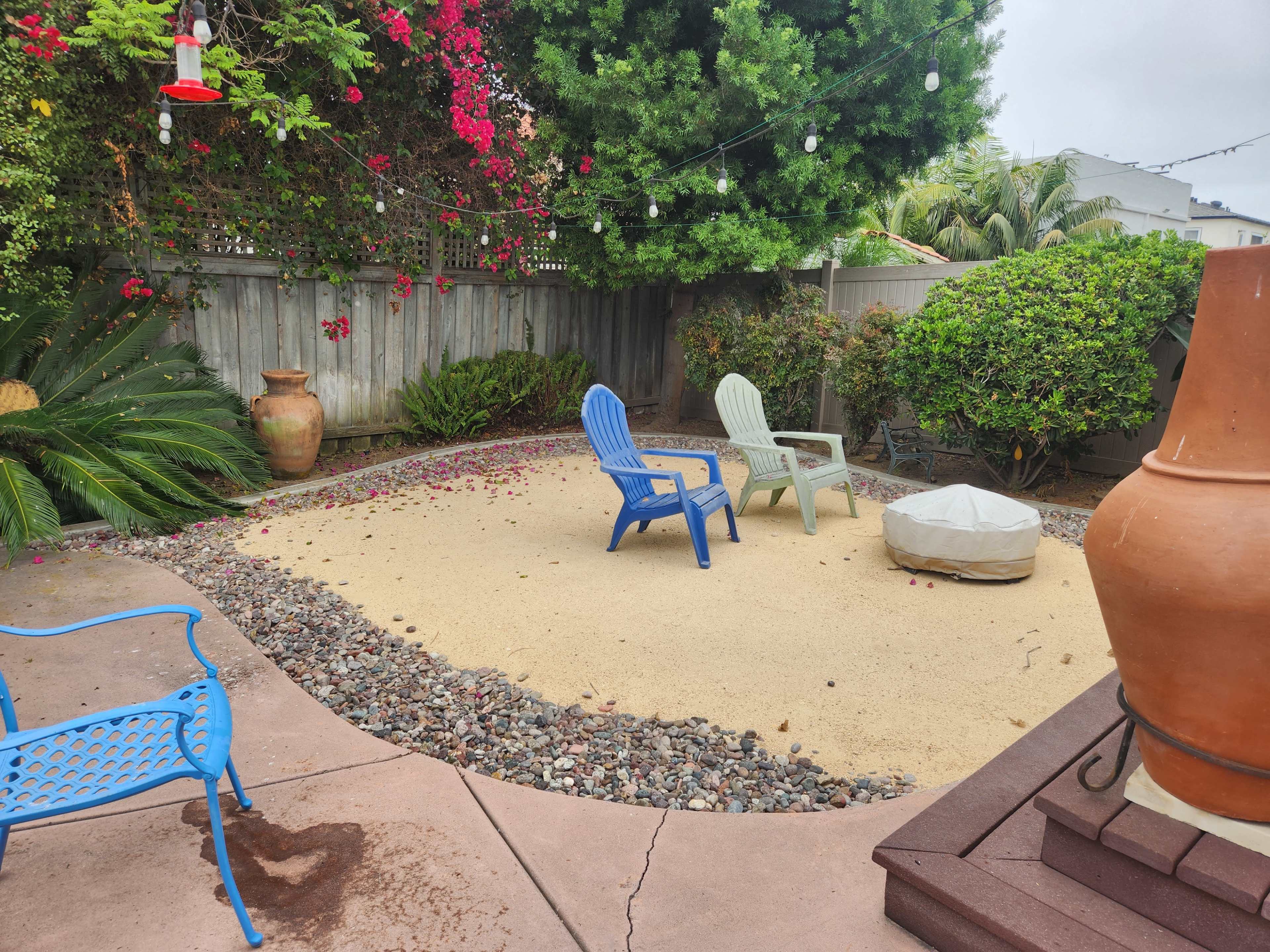 The image shows a small outdoor patio area with two colorful chairs and a covered fire pit surrounded by rocks and greenery.