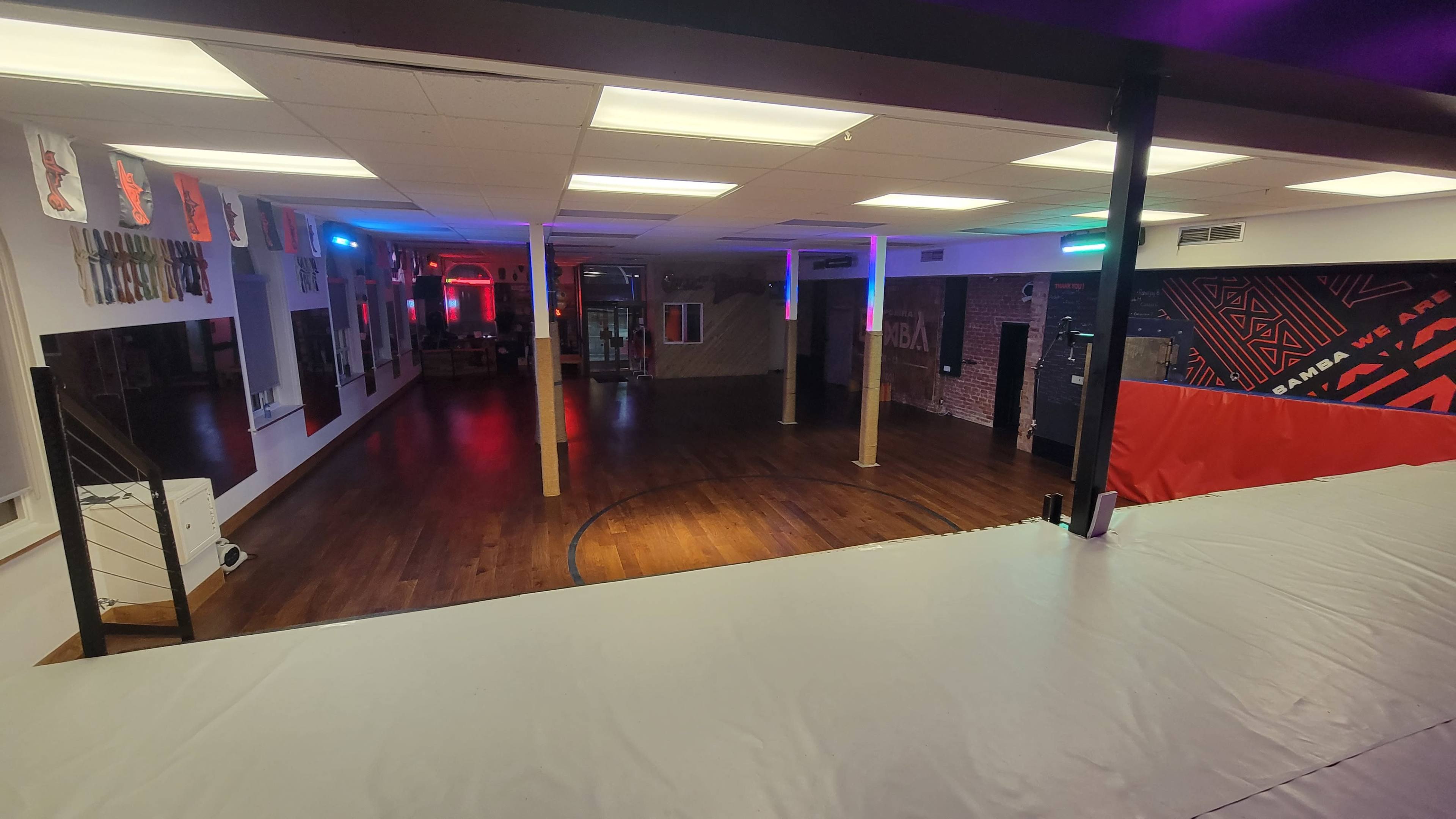 The image shows a spacious training facility with wooden flooring, brightly colored lights, and various martial arts equipment arranged along the walls.