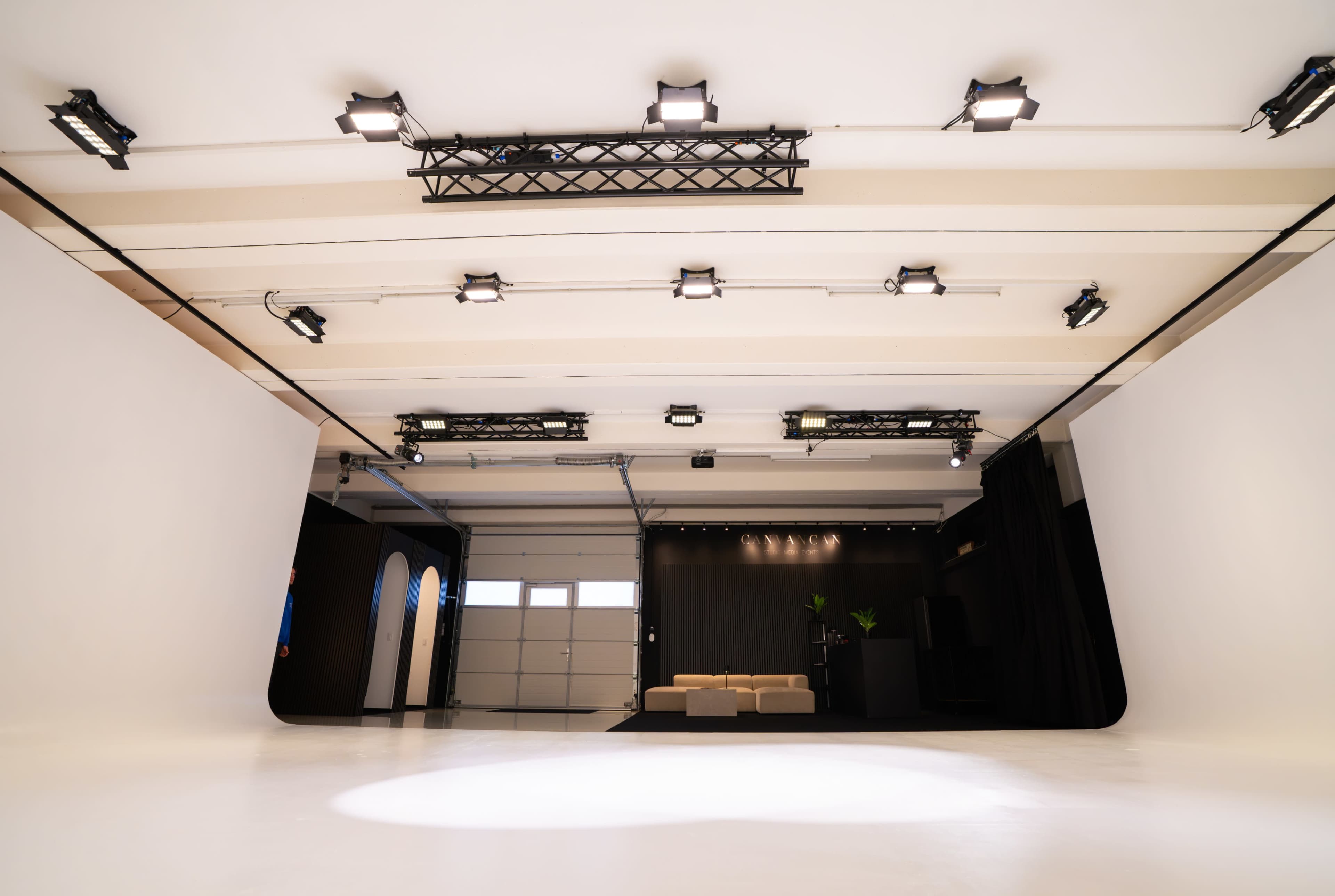 The image shows a spacious photography or video studio with a white floor, multiple overhead lights, and a minimalist black backdrop featuring a sofa and indoor plants.