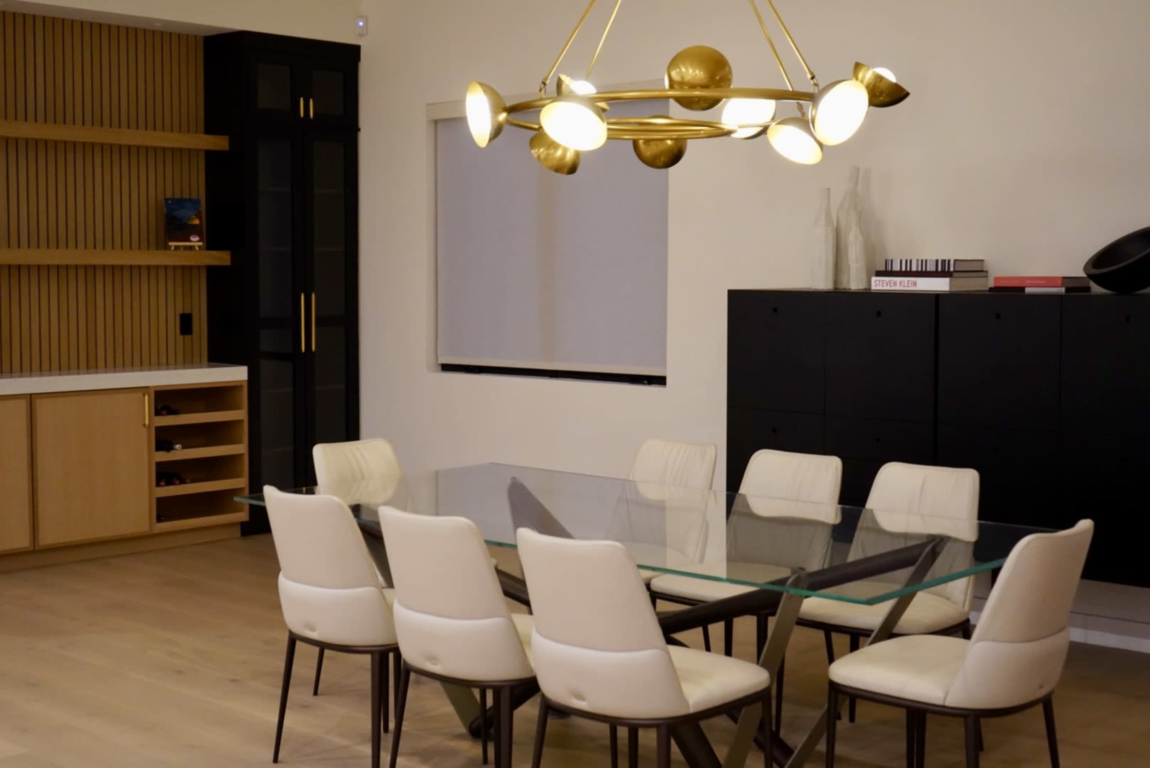 A modern dining area features a large glass table surrounded by eight cream-colored chairs, with a stylish light fixture overhead and a wooden accent wall in the background.