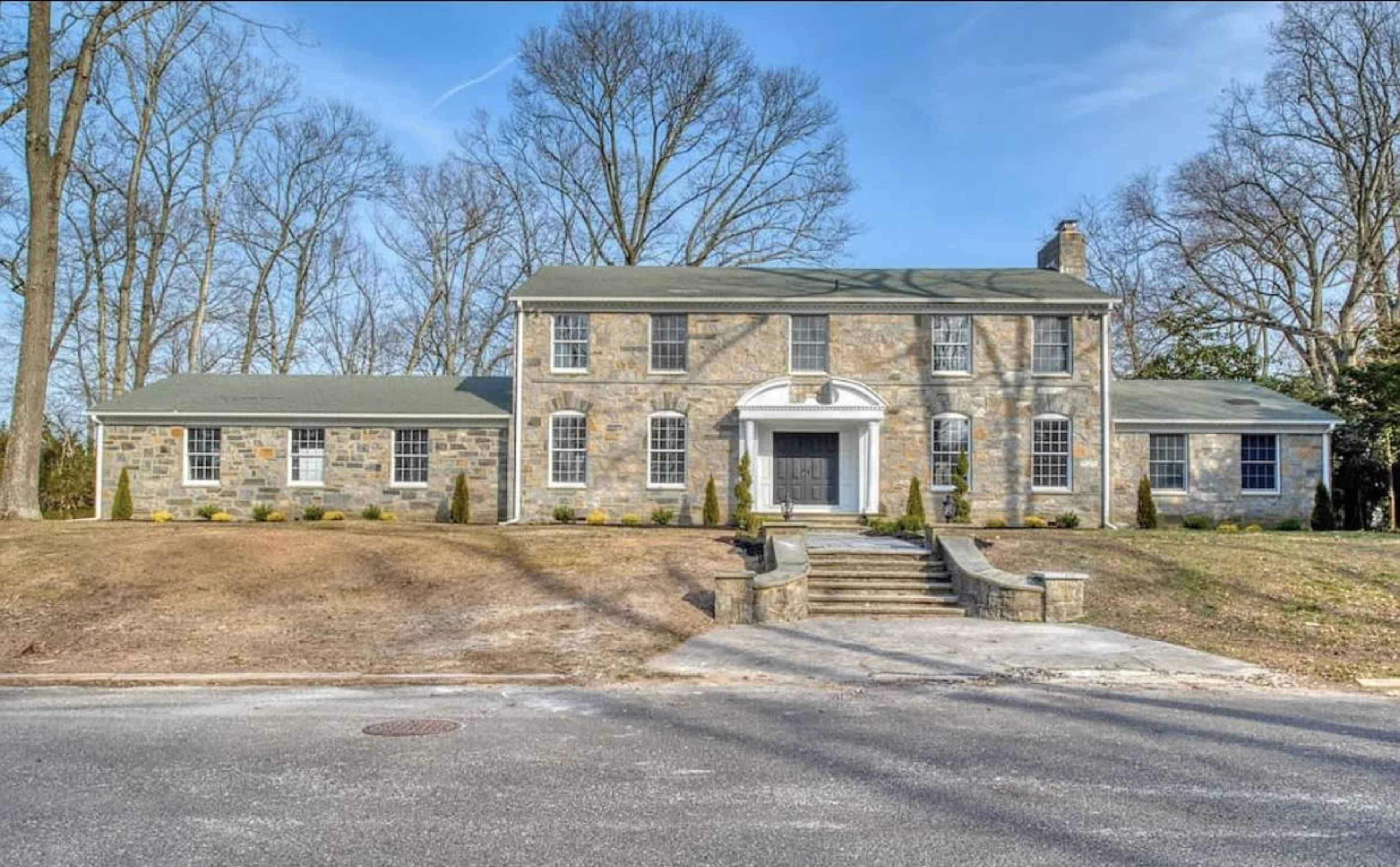 A large, two-story stone house with a symmetrical facade and a central entrance, surrounded by bare trees and a grassy lawn.