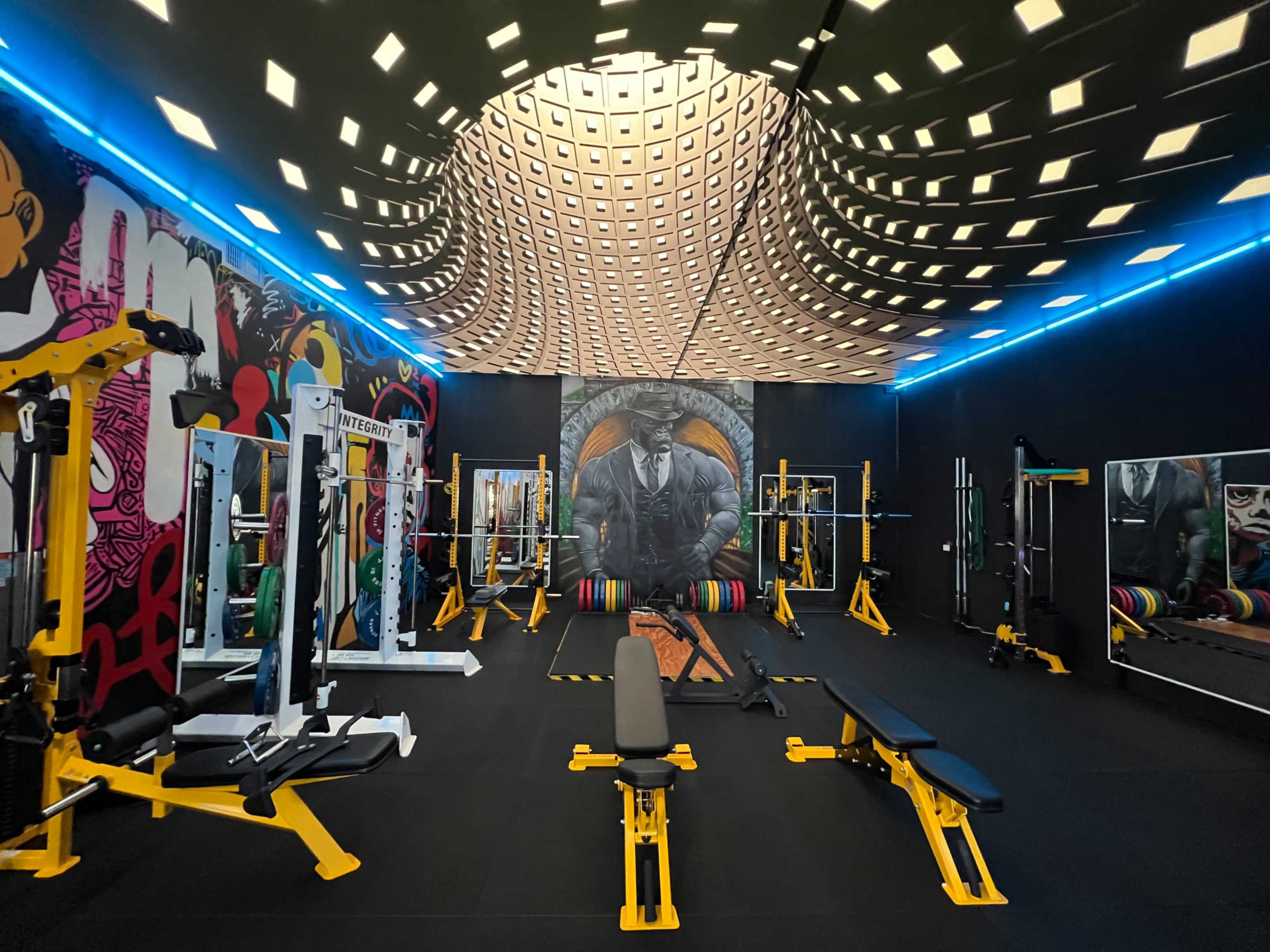 A modern gym interior featuring various weightlifting equipment, colorful wall art, and a distinctive patterned ceiling.