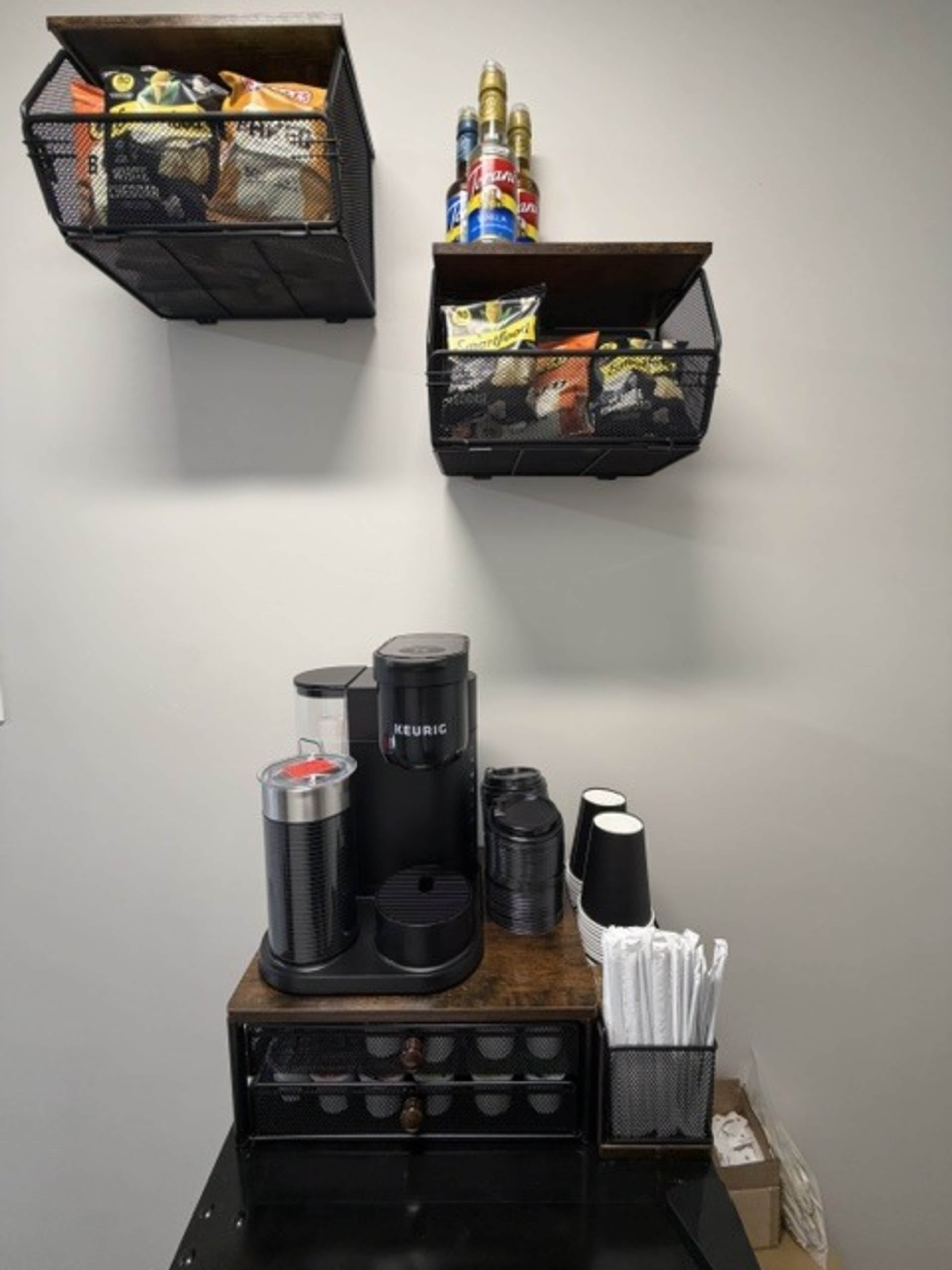 The image shows a coffee station with a Keurig machine, disposable cups, and two wall-mounted baskets filled with snacks and condiments.