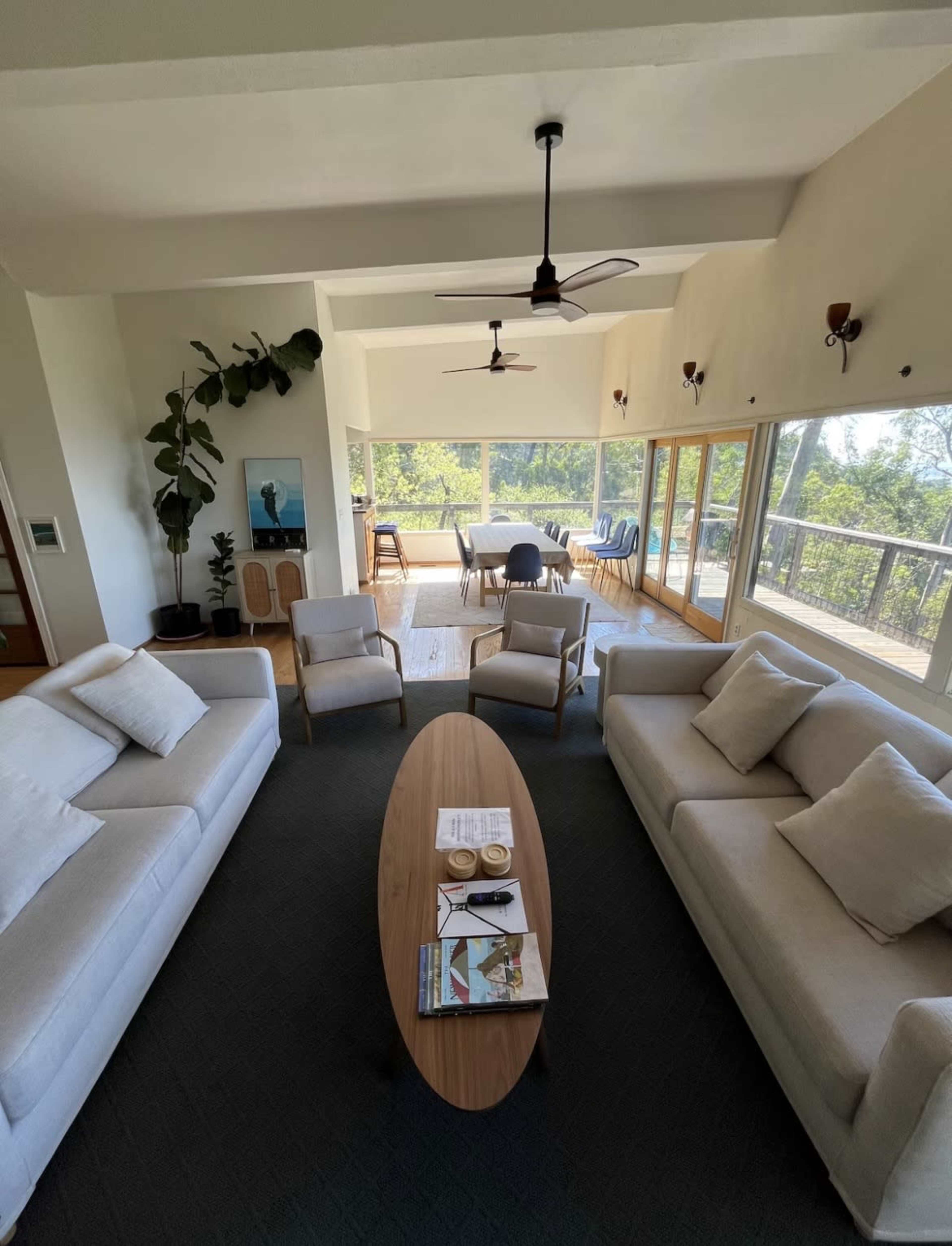 A spacious living room features two light-colored sofas, a wooden coffee table, and large windows overlooking a verdant outdoor area.
