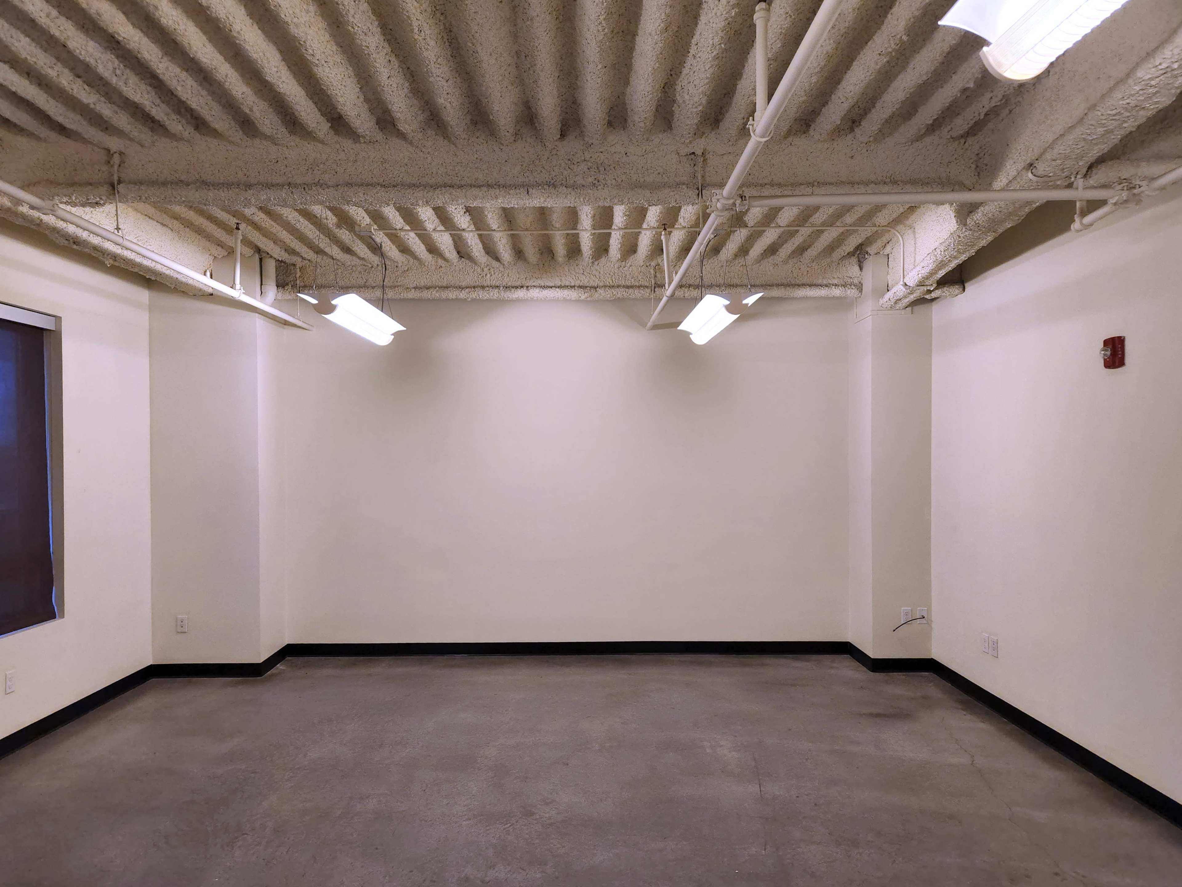 An empty room with a concrete floor, white walls, and fluorescent lighting along the ceiling.