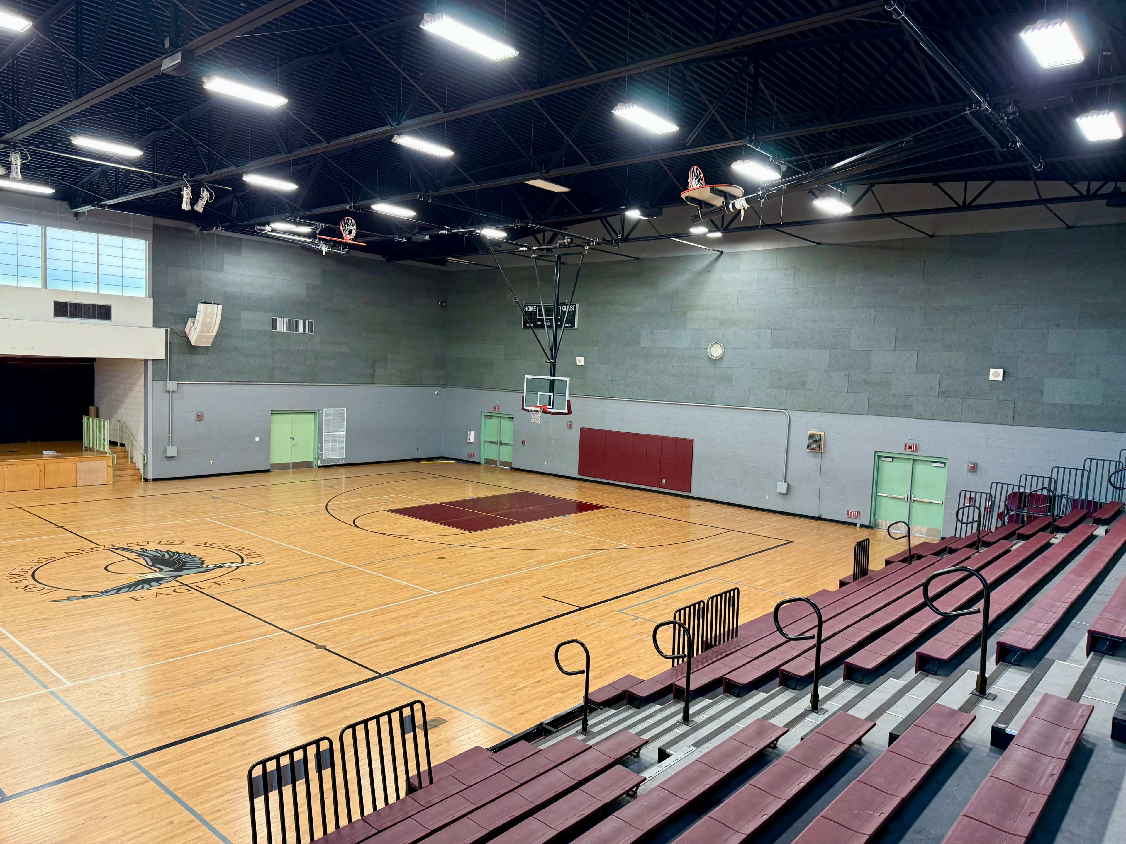 The image shows a spacious wooden gymnasium with bleachers, basketball hoops, and a polished court.