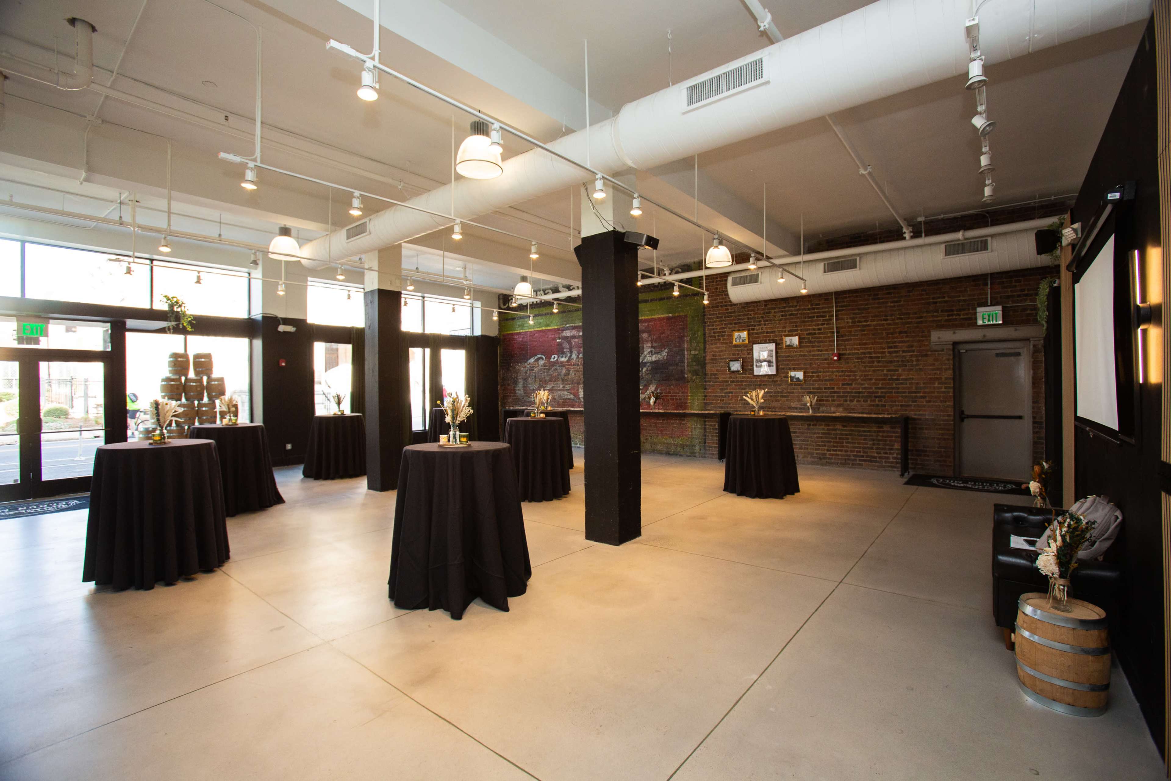 The image shows a spacious event venue with a polished concrete floor, black tablecloths arranged at various points, and brick walls adorned with artwork.