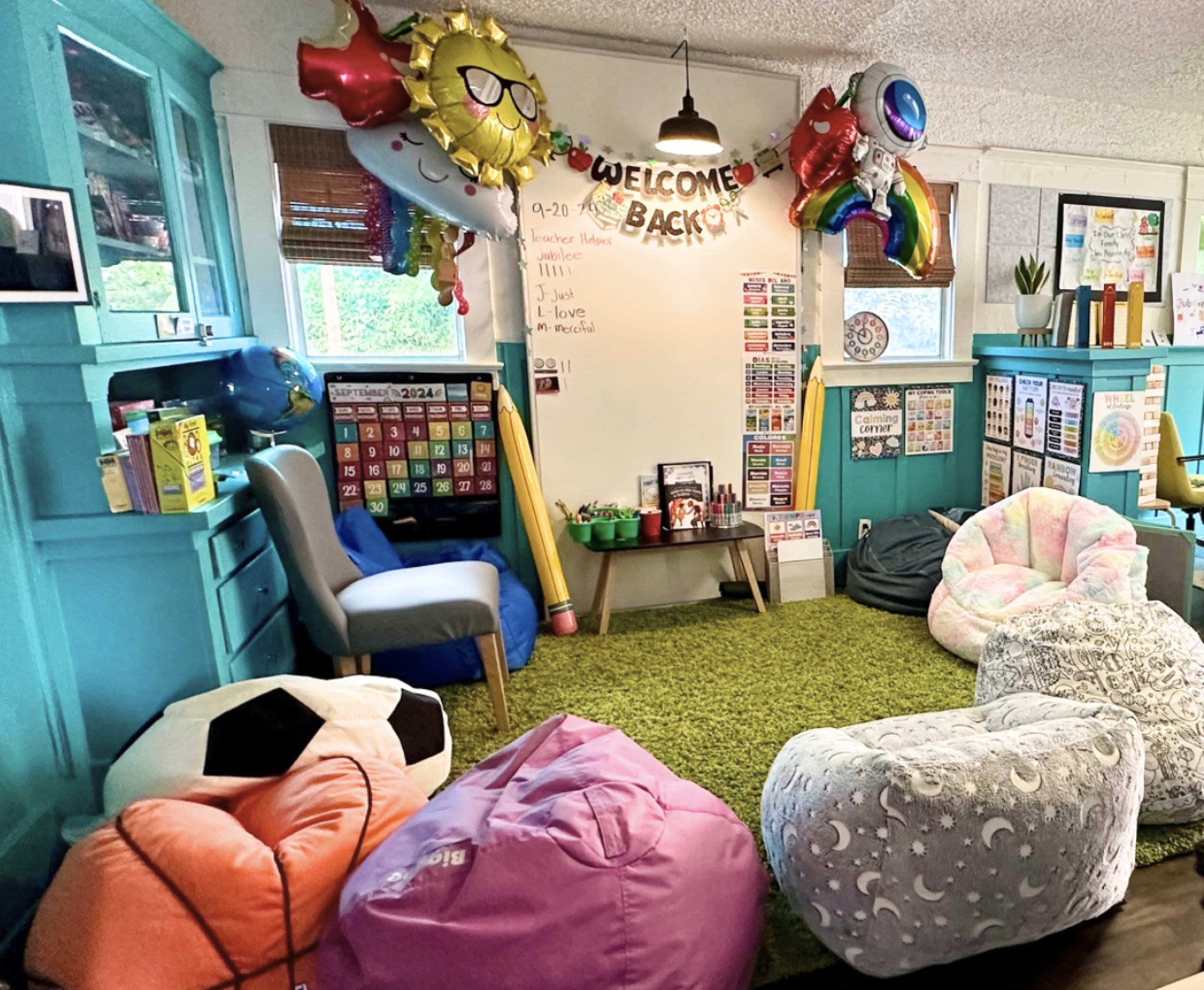 The image shows a colorful classroom area with bean bag chairs, a green carpet, a whiteboard displaying "WELCOME BACK," and various educational posters and decorations on the walls.