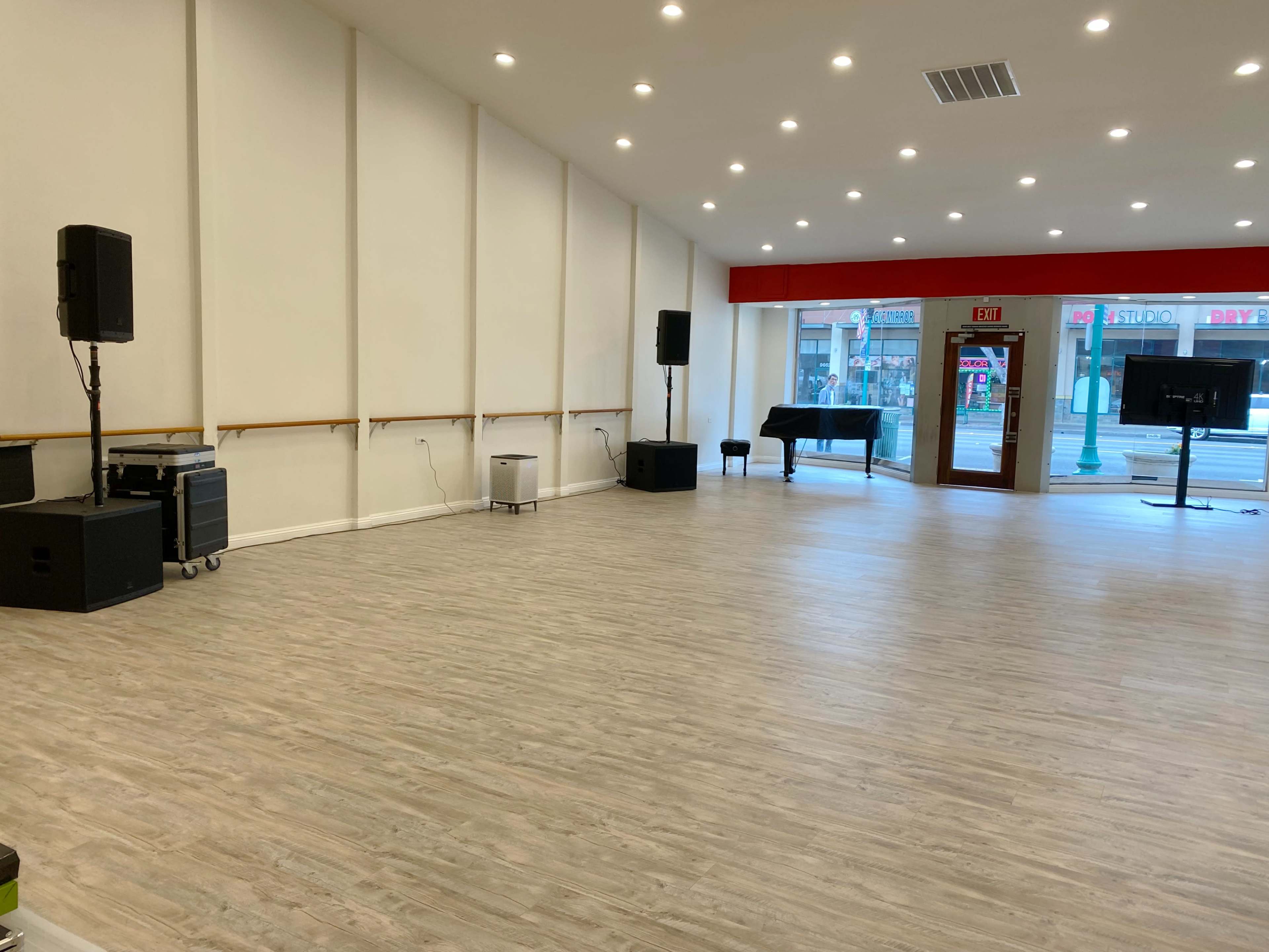 A spacious, empty room with hardwood flooring, equipped with audio speakers, a piano, and two screens against the walls.