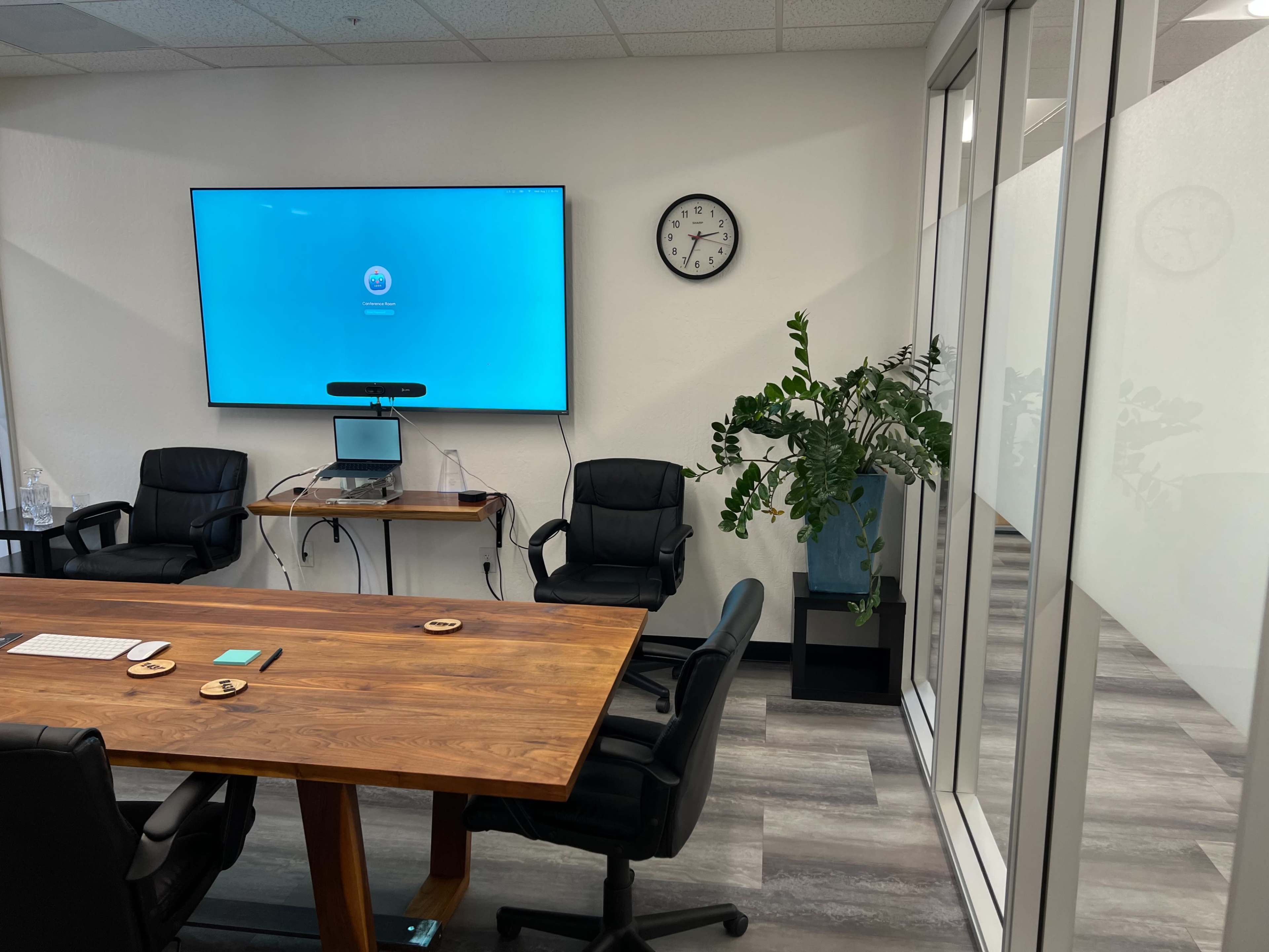 A conference room with a large screen on the wall, a clock, a wooden table surrounded by black chairs, and a potted plant in the corner.