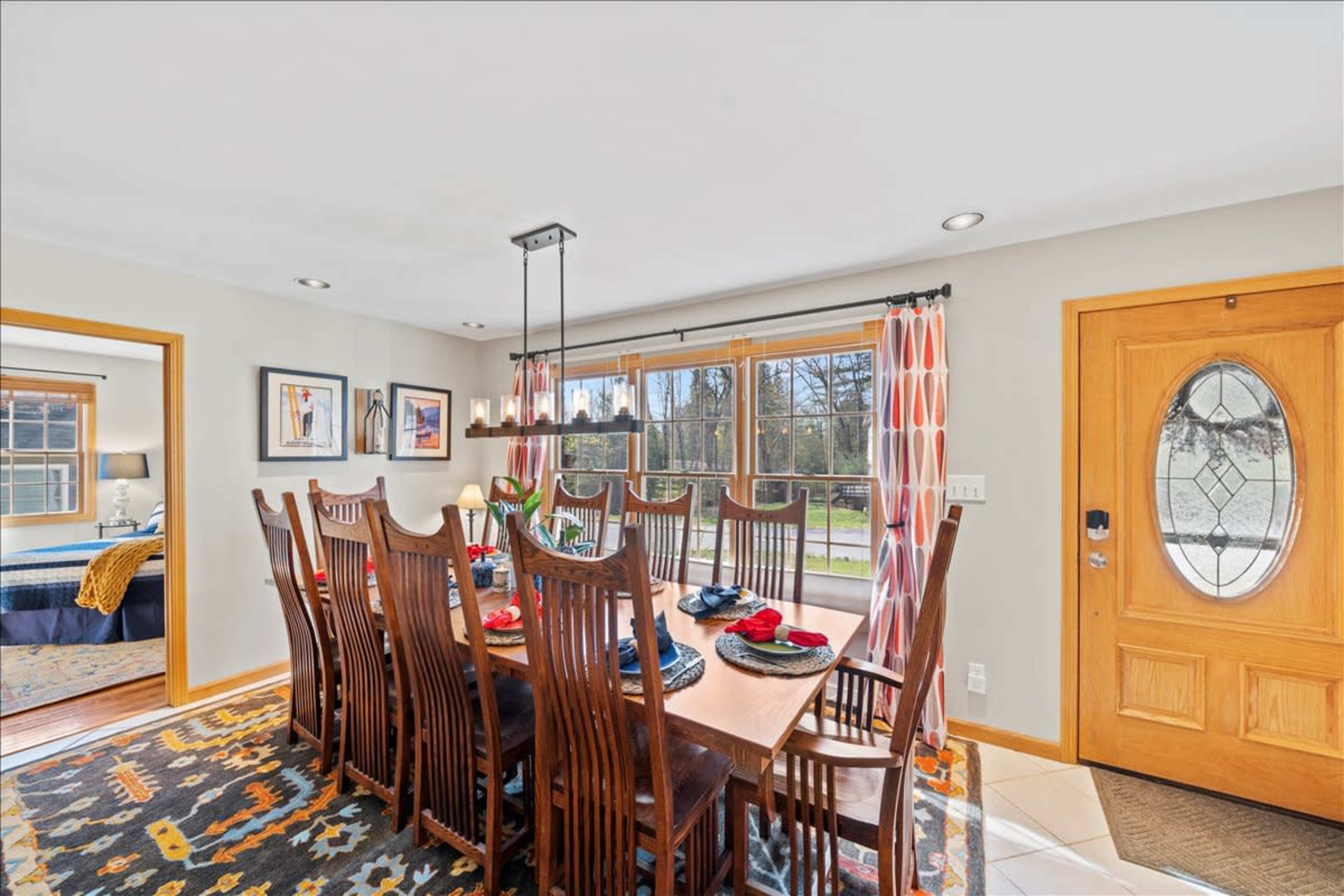 A spacious dining room features a large wooden table set with colorful place settings, flanked by tall windows displaying a view of the outdoors.