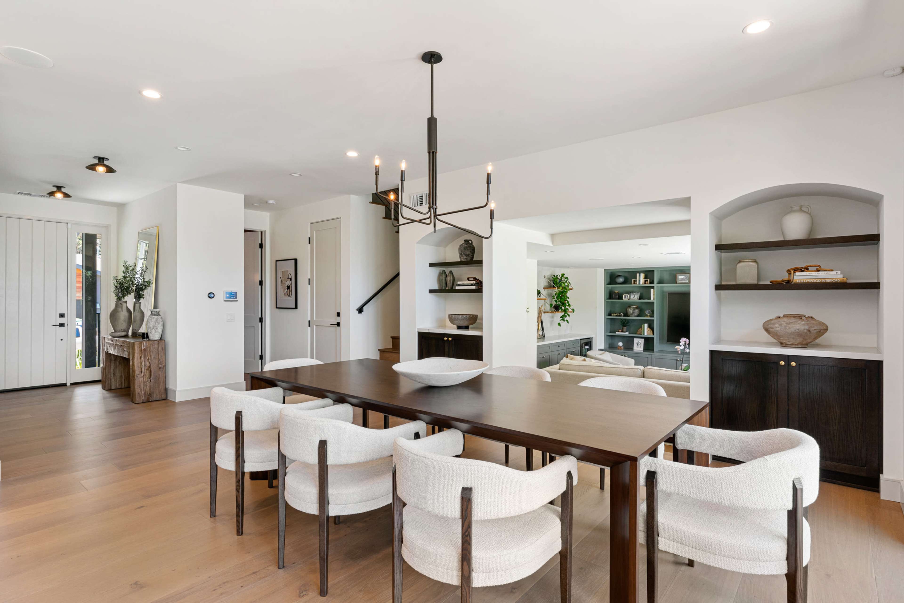 A modern dining area features a wooden table surrounded by upholstered chairs, with a decorative chandelier overhead and access to adjacent rooms.