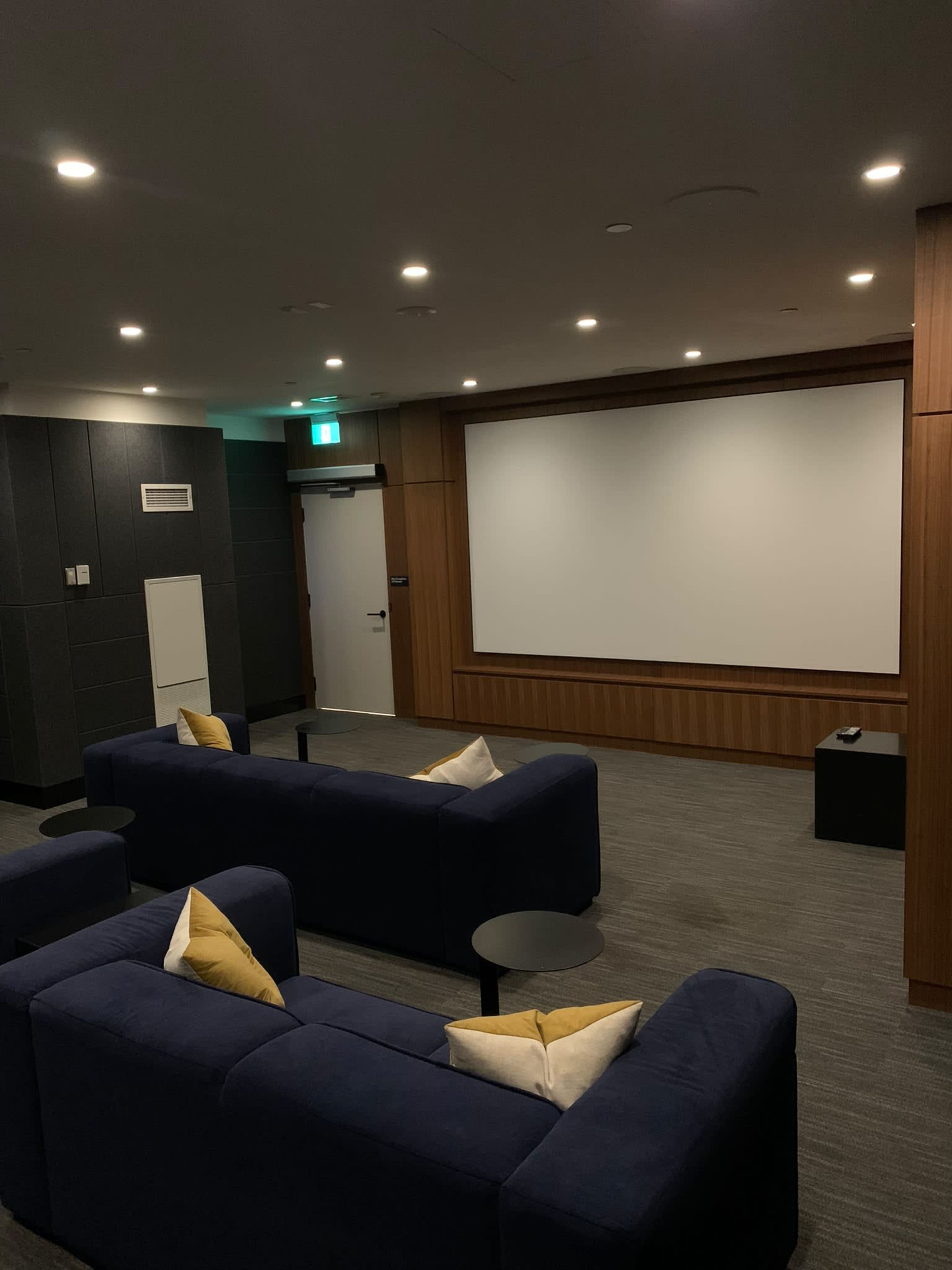 The image shows a small, modern screening room furnished with blue sofas and a large blank projection screen on the wall.