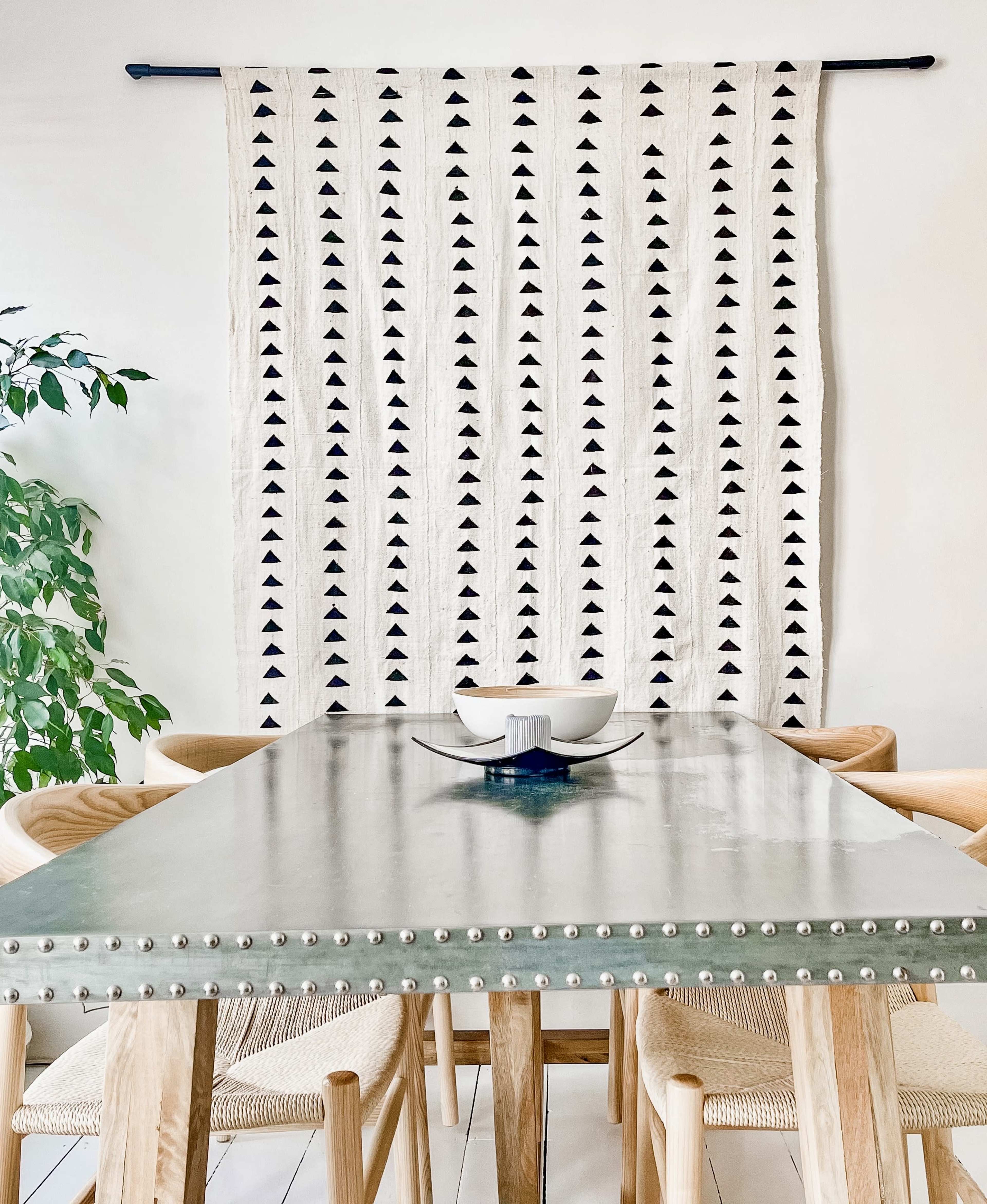 A dining table with a metal surface and studded edges is set beneath a patterned wall hanging and alongside a potted plant.
