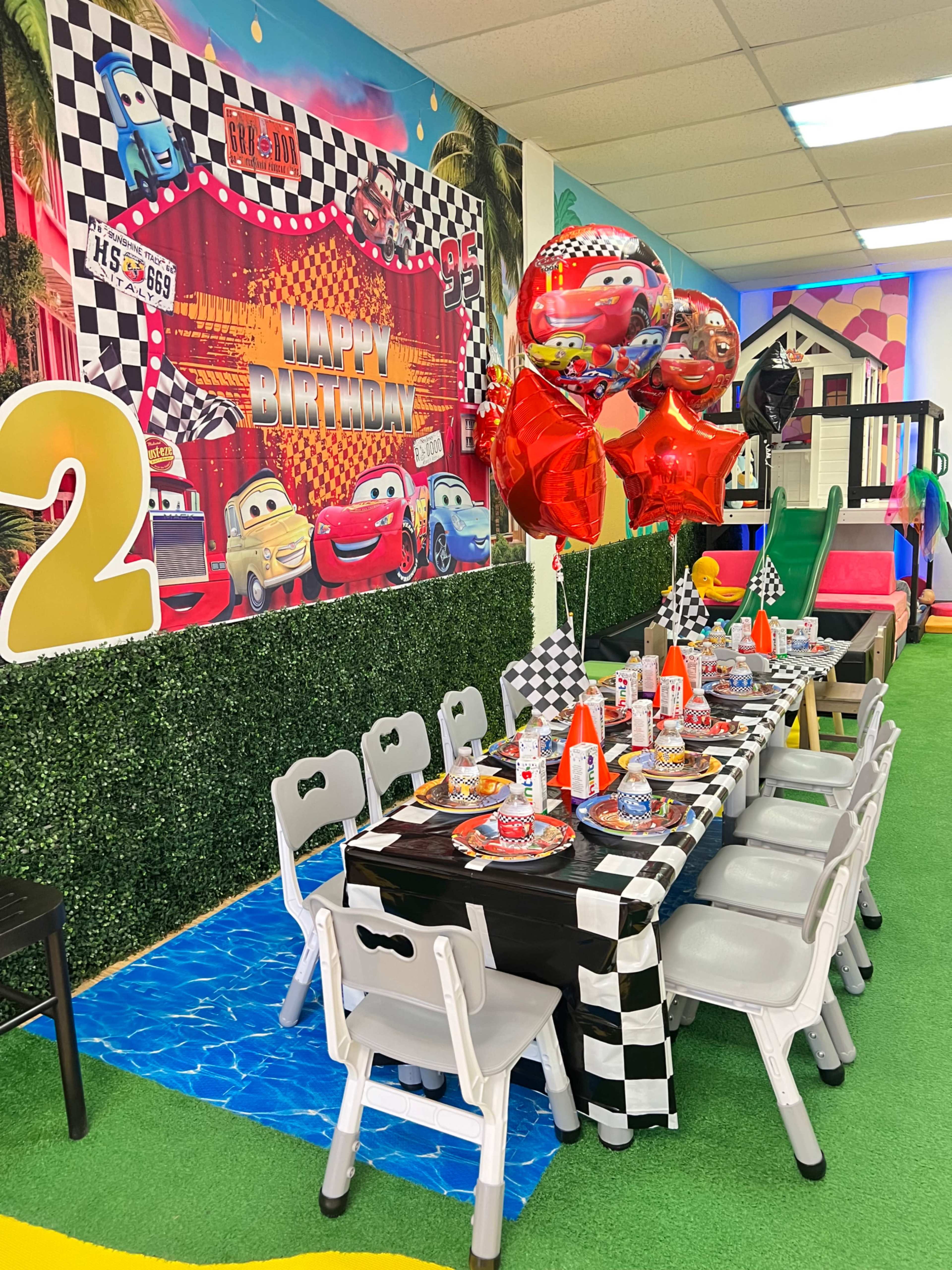 A birthday party setup features a checkered tablecloth with themed decorations, balloons, and place settings arranged around a long table in a colorful playroom.