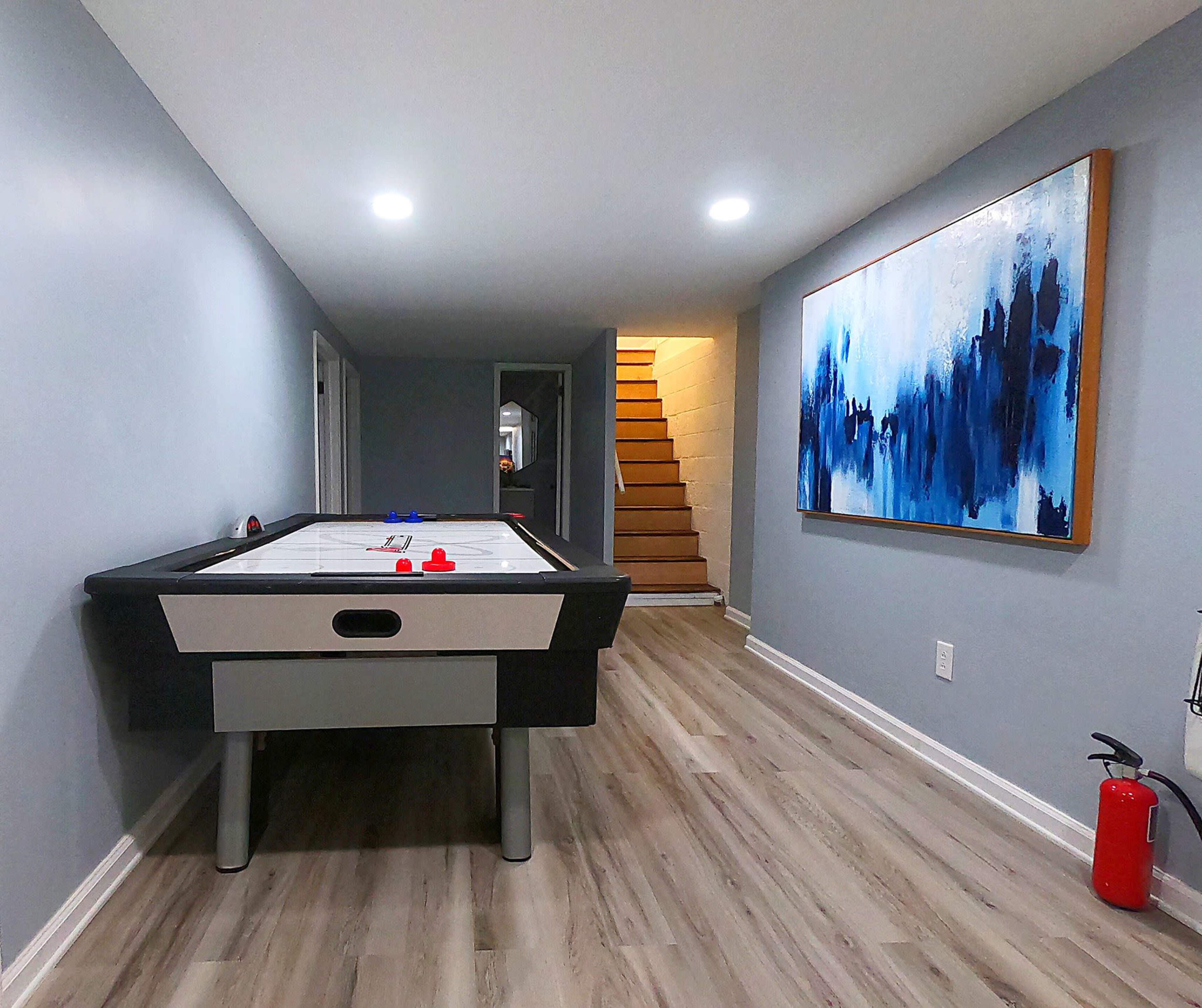 A game room features an air hockey table, a large abstract painting on the wall, and a staircase in the background.
