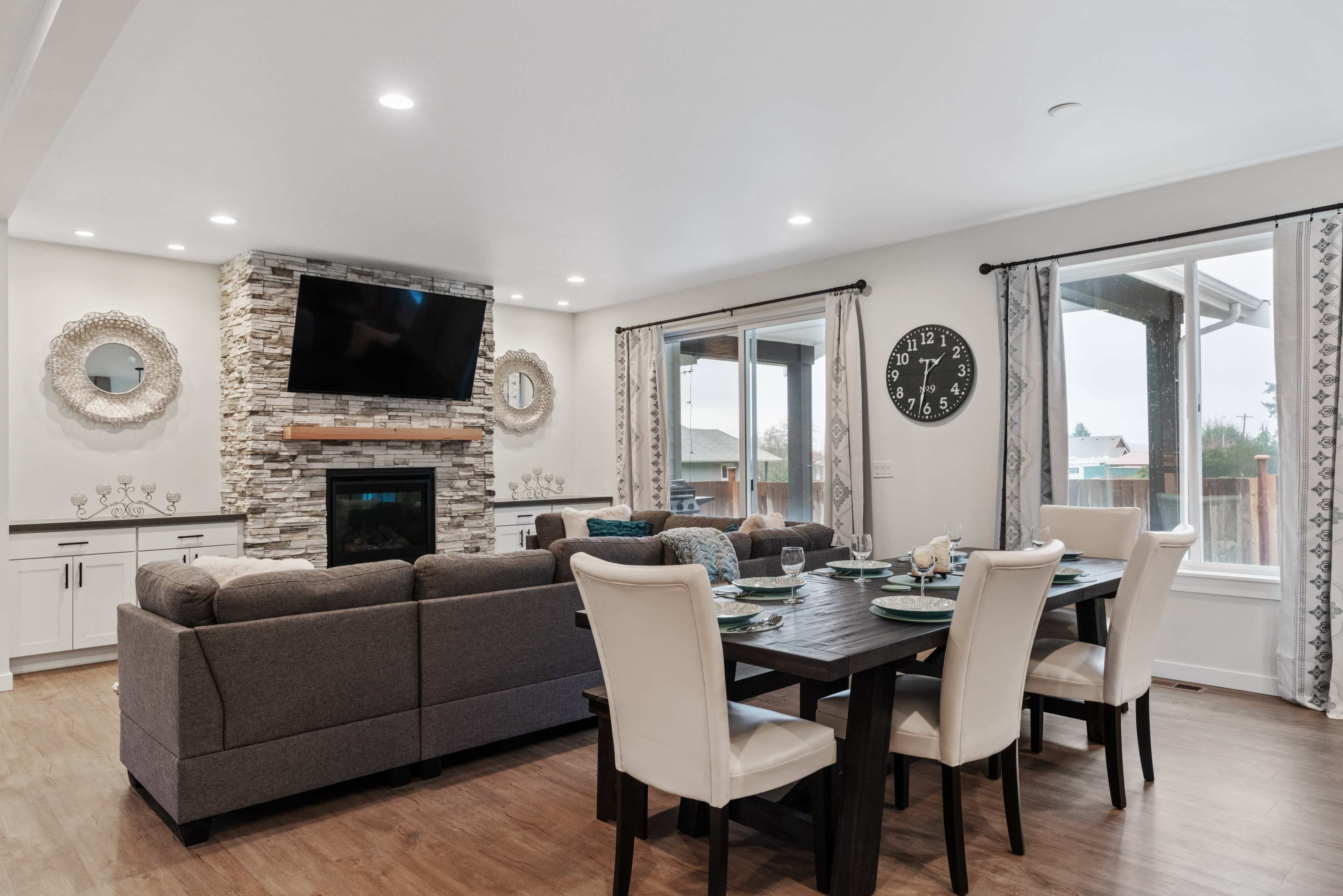 A modern living room features a stone fireplace with a mounted TV, surrounded by a dining area with a dark wooden table set for meals.