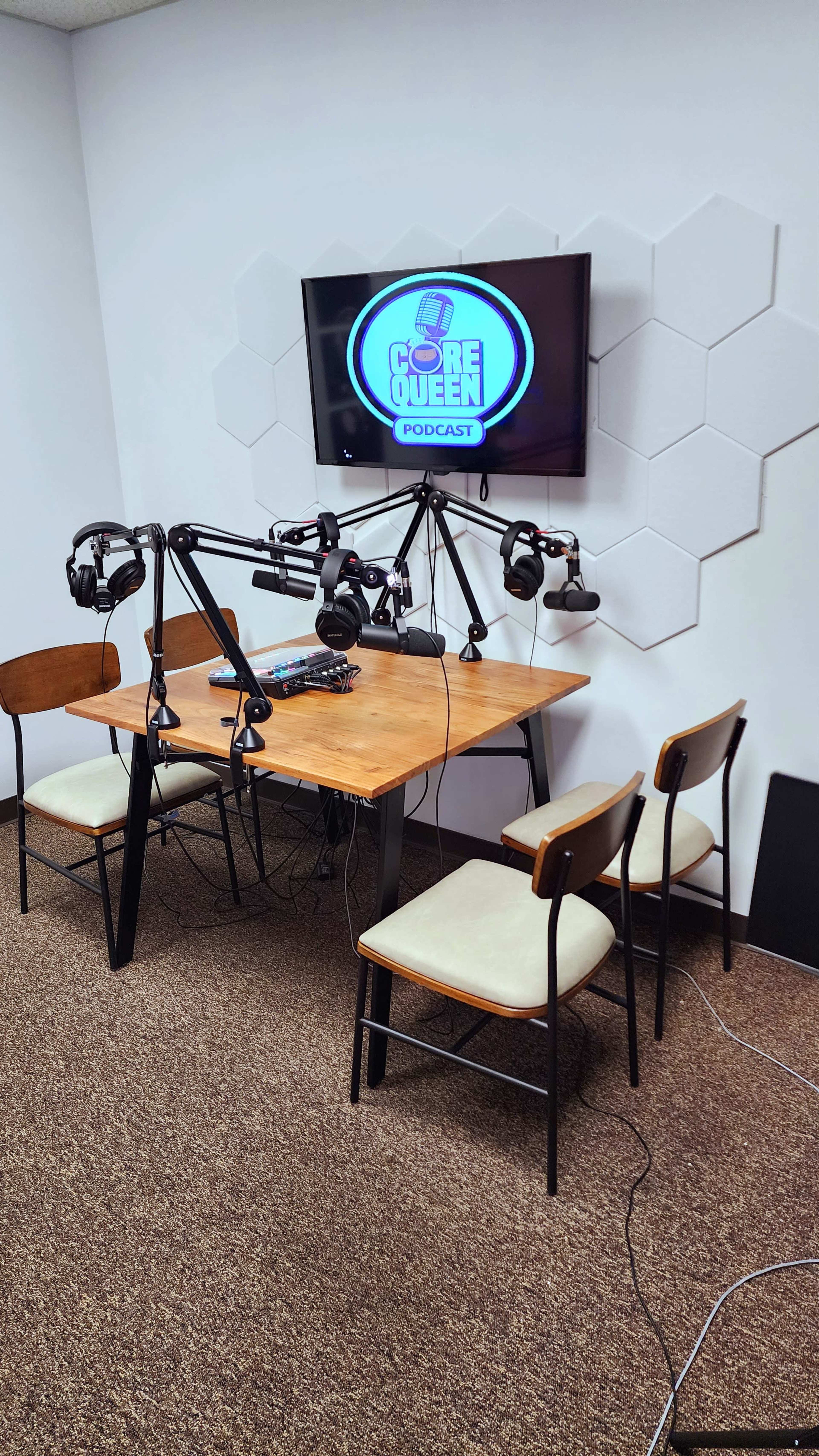 A podcast recording studio features a wooden table surrounded by four chairs, with microphones and headphones mounted on adjustable arms, and a television displaying the "Core Queen Podcast" logo.