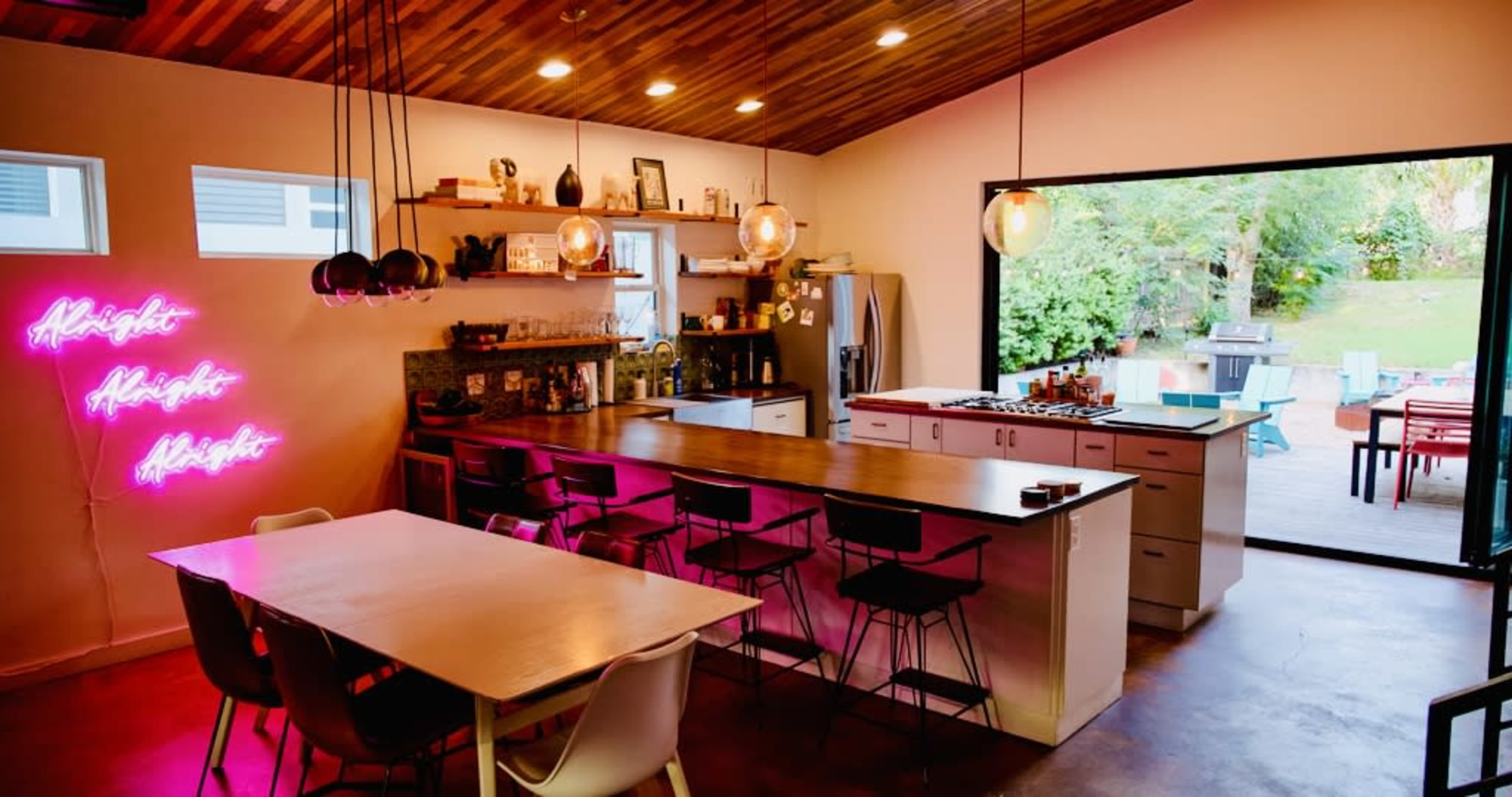 The image shows a modern kitchen and dining area featuring a large island with bar stools, a neon sign, and an open wall leading to an outdoor patio filled with furniture.