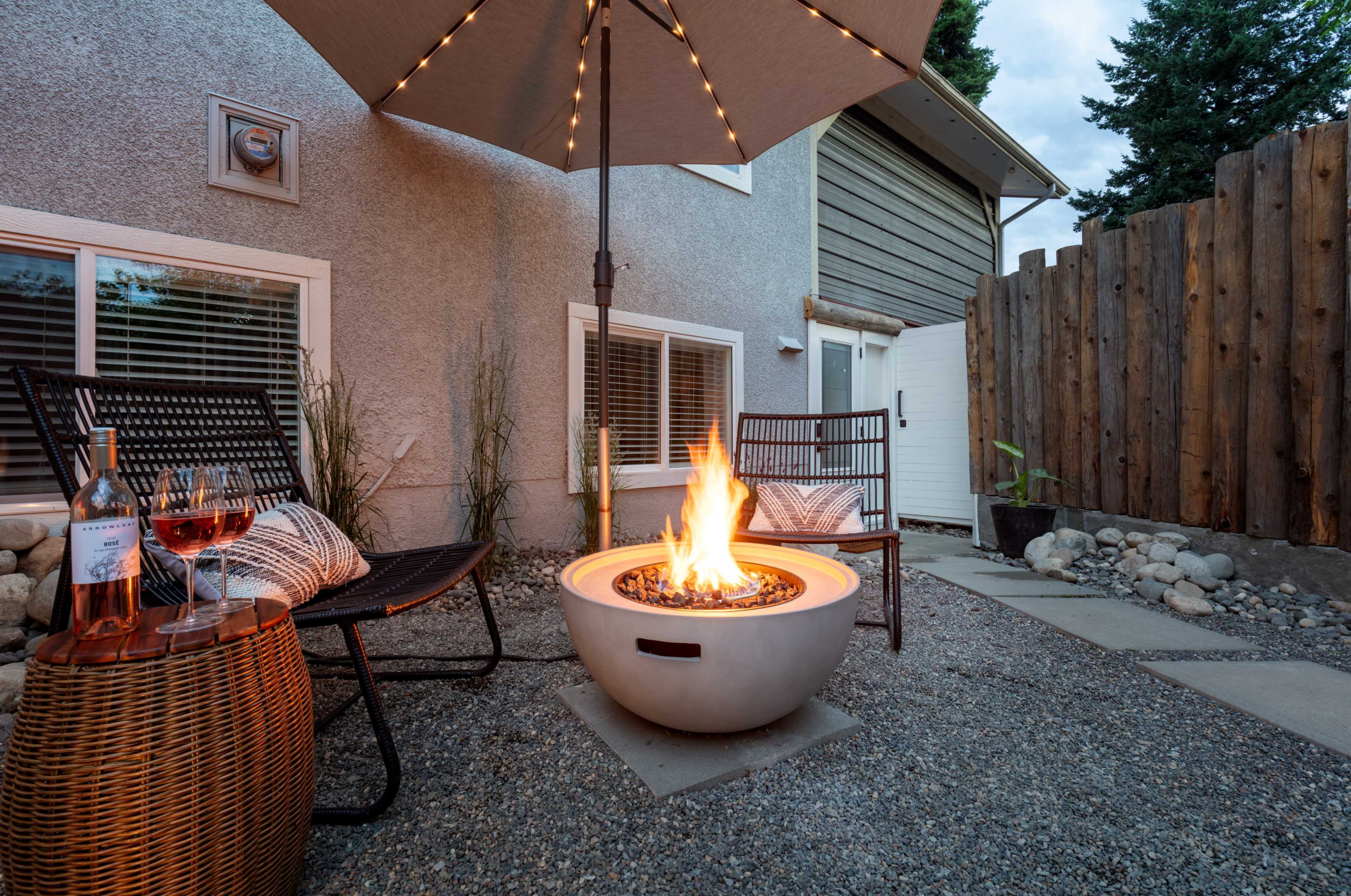 A modern outdoor seating area features two chairs and a round fire pit with flames, surrounded by gravel and a stone wall.