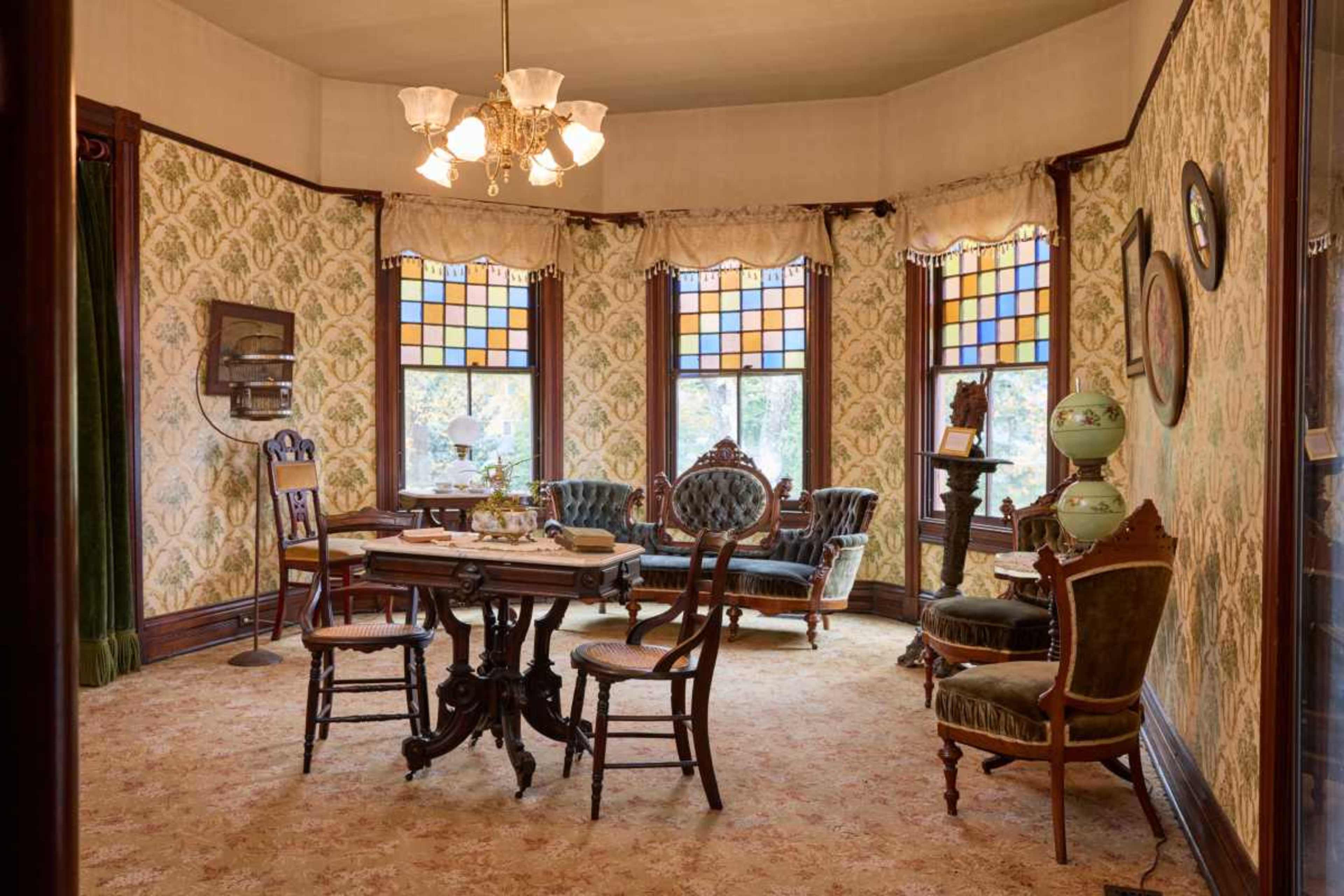 The image shows a vintage parlor room with patterned wallpaper, ornate furniture, and colorful stained glass windows.