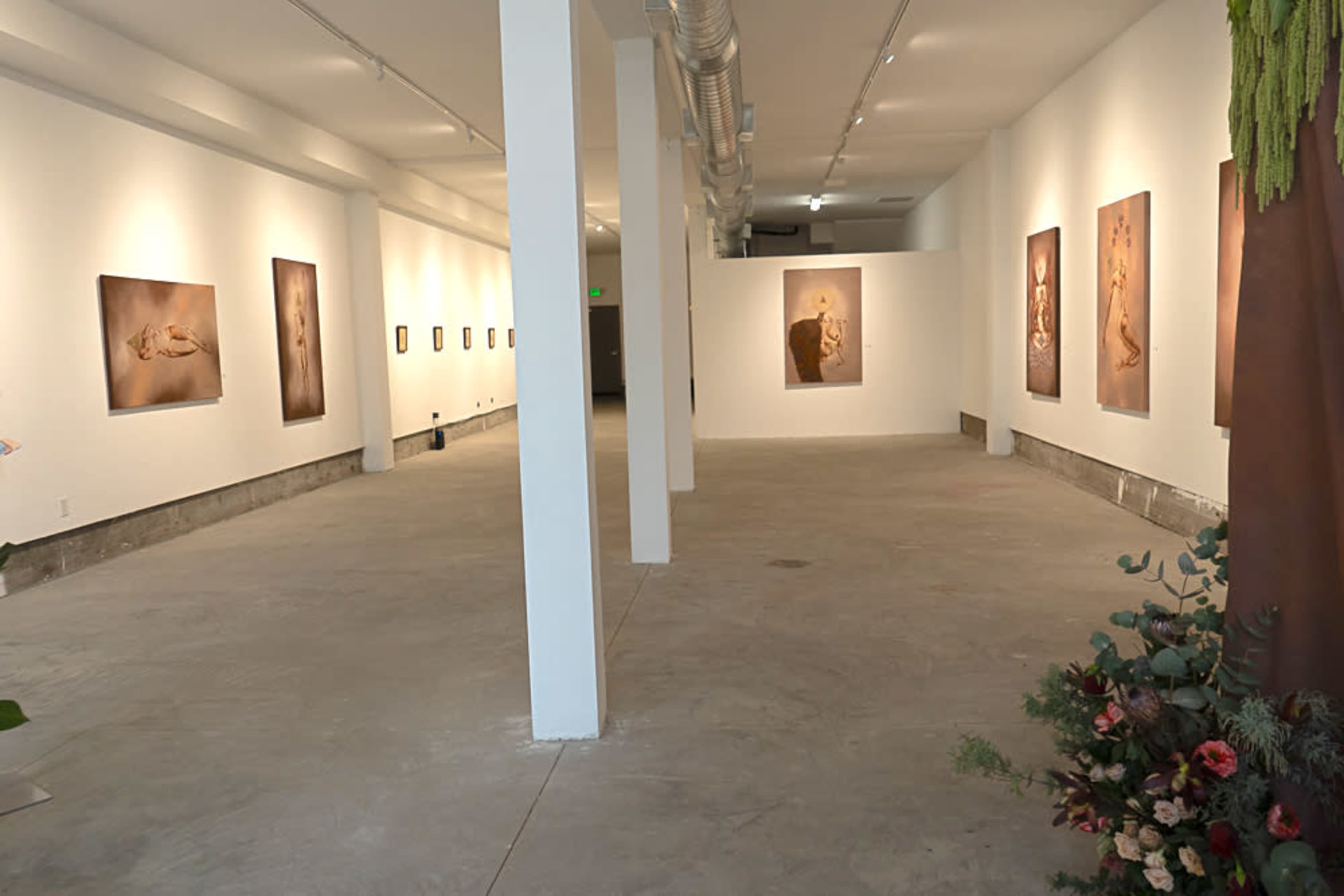 The image shows a spacious art gallery with white walls displaying several framed artworks, accompanied by a concrete floor and minimal decorations.
