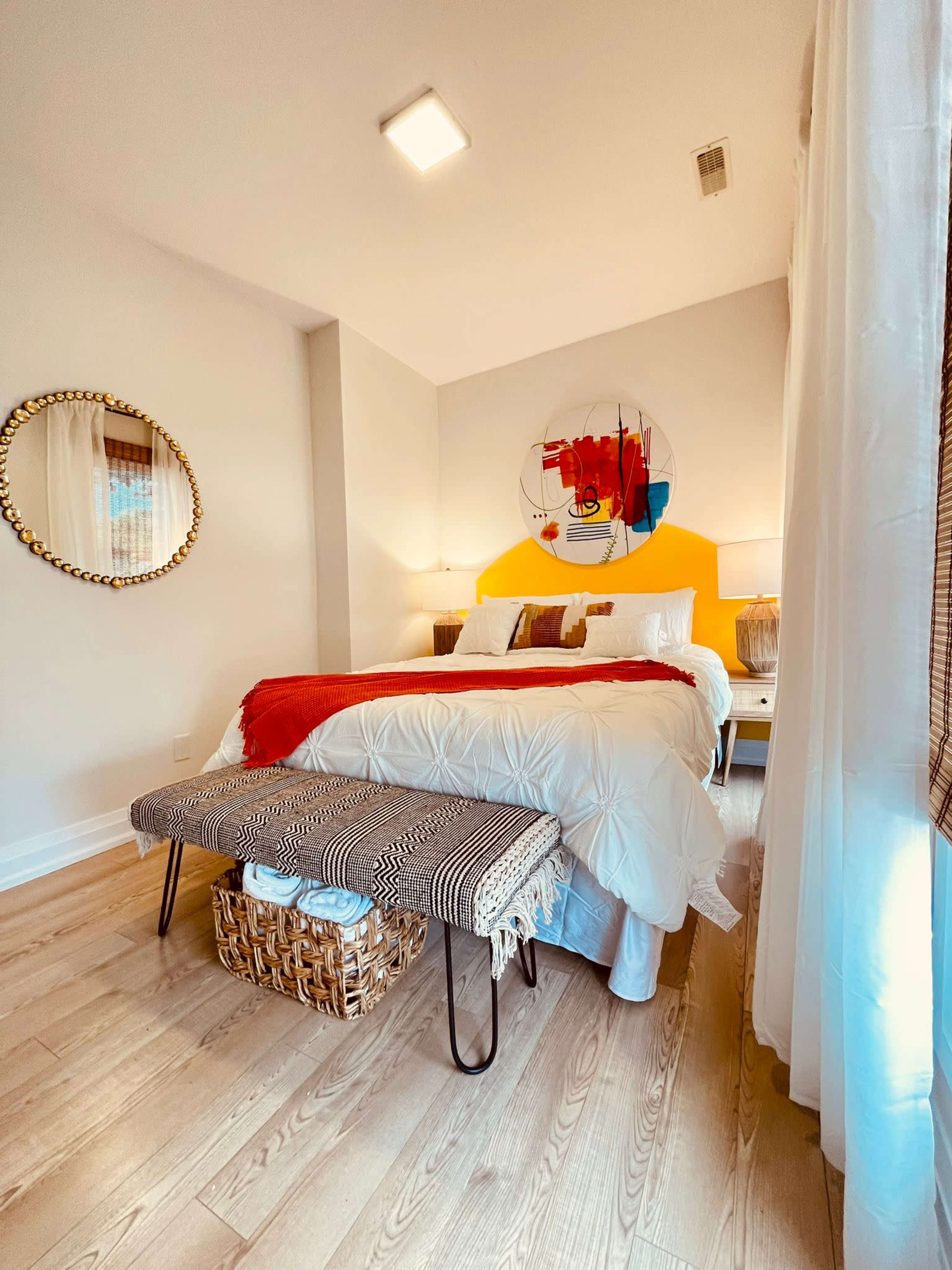 A neatly arranged bedroom featuring a large bed with white bedding, a colorful abstract art piece on the wall, and a woven storage basket at the foot of the bed.