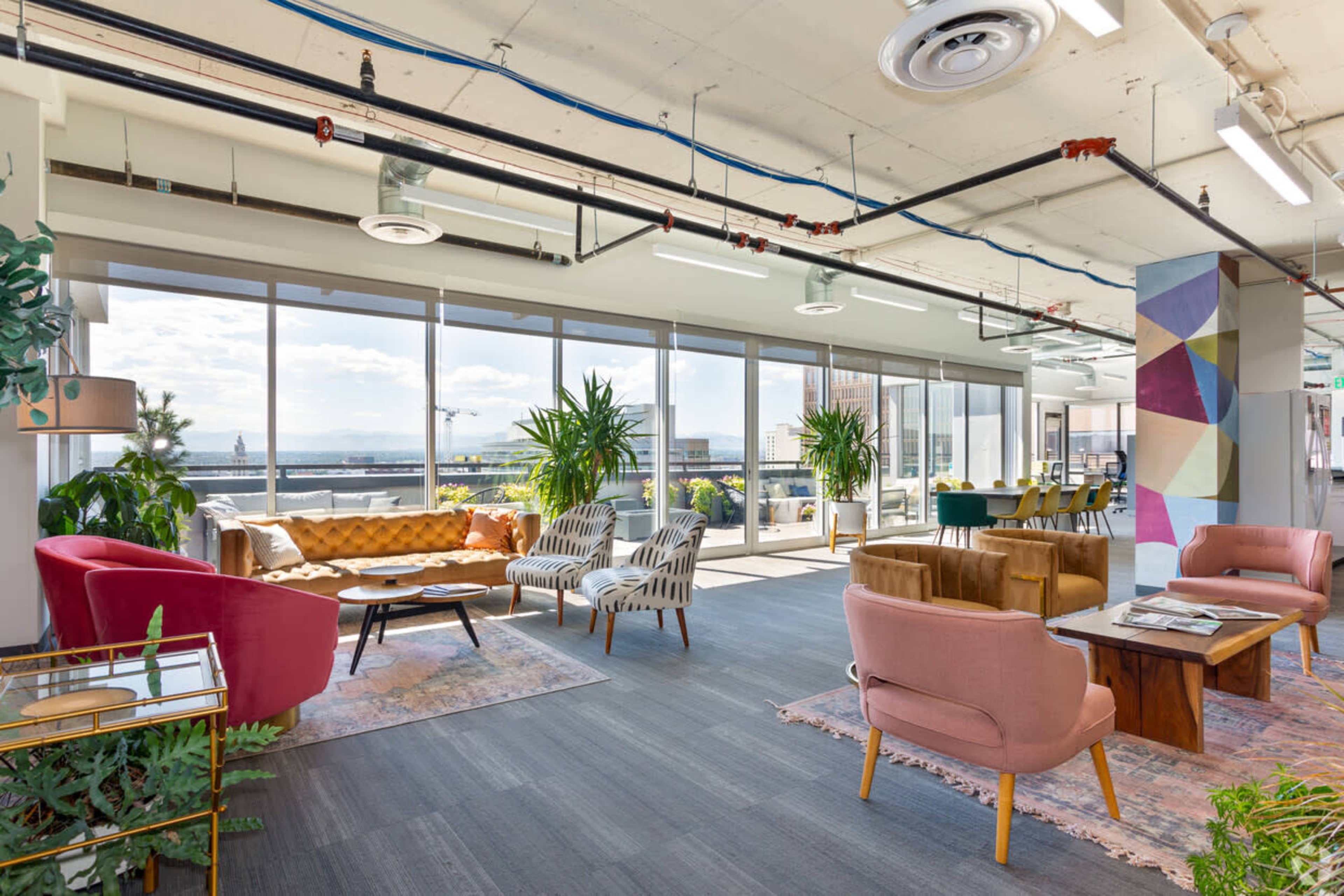 A modern office lounge area with various seating arrangements, large windows, and an open layout featuring plants and decorative rugs.