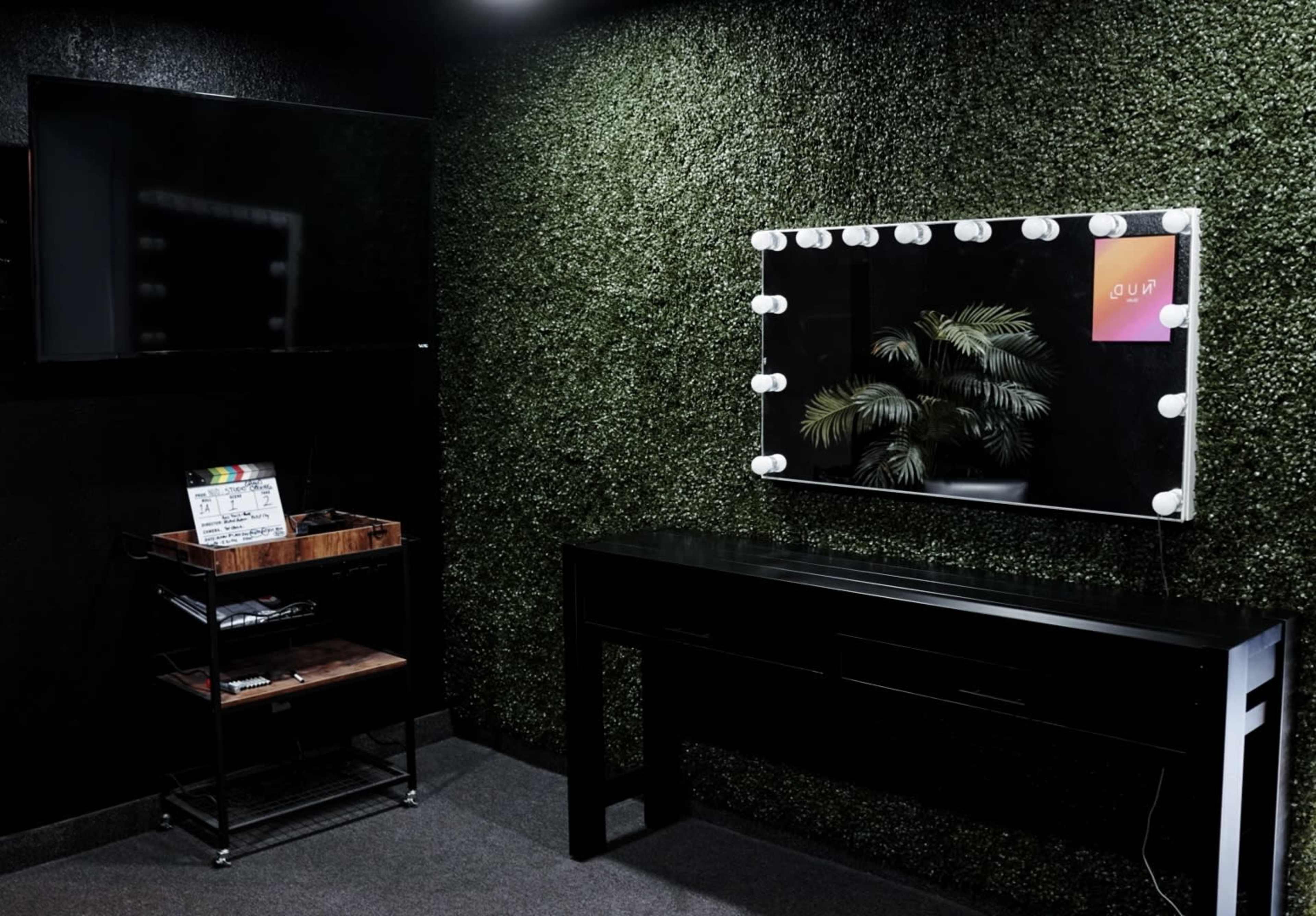 A room with a wall of artificial greenery, a black console table beneath a mirror framed with lights, and a television mounted on the wall.