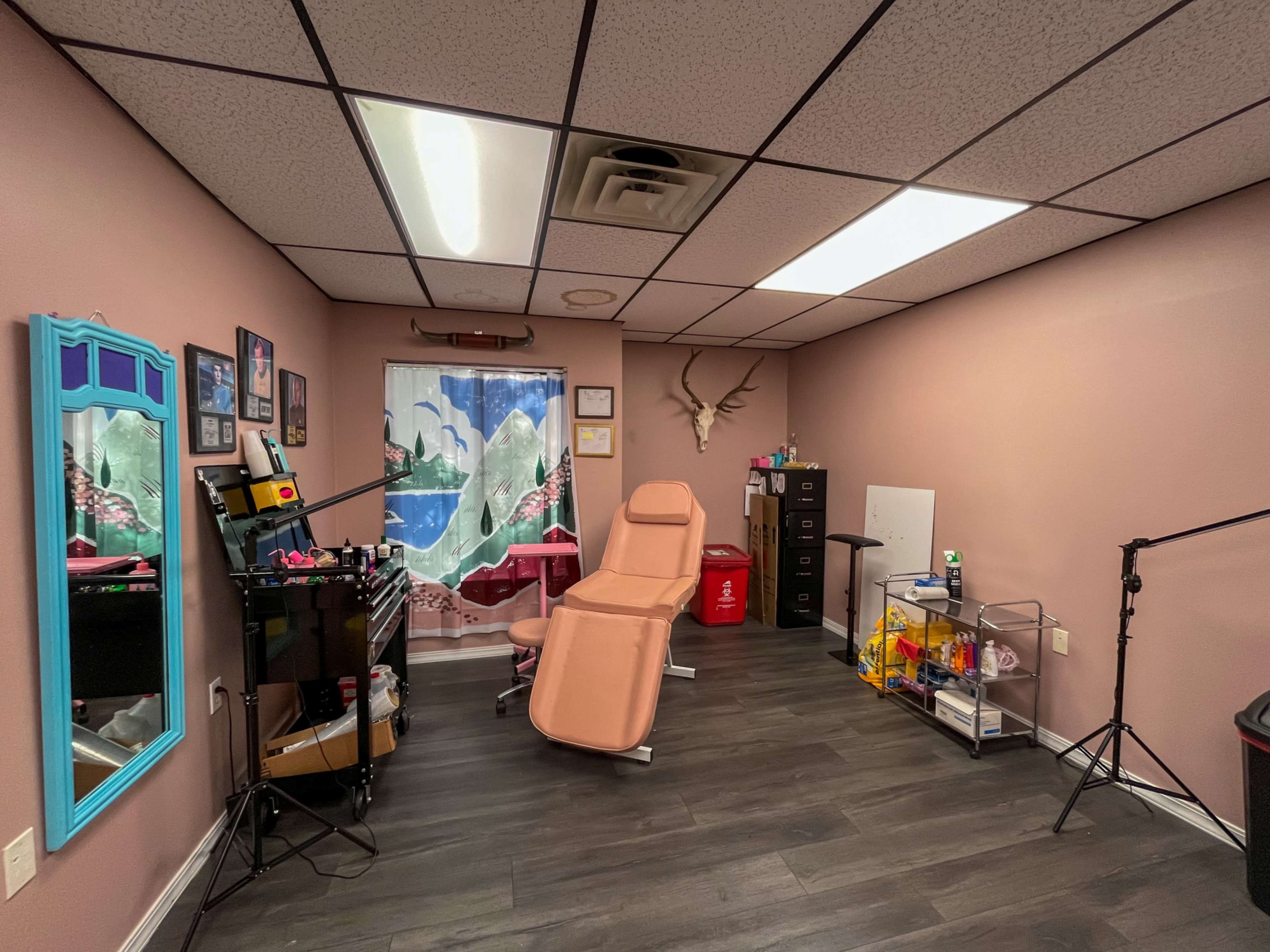 The image shows a small, inviting room designed for beauty or tattoo services, featuring a treatment chair, various equipment on a workstation, and a light source overhead.