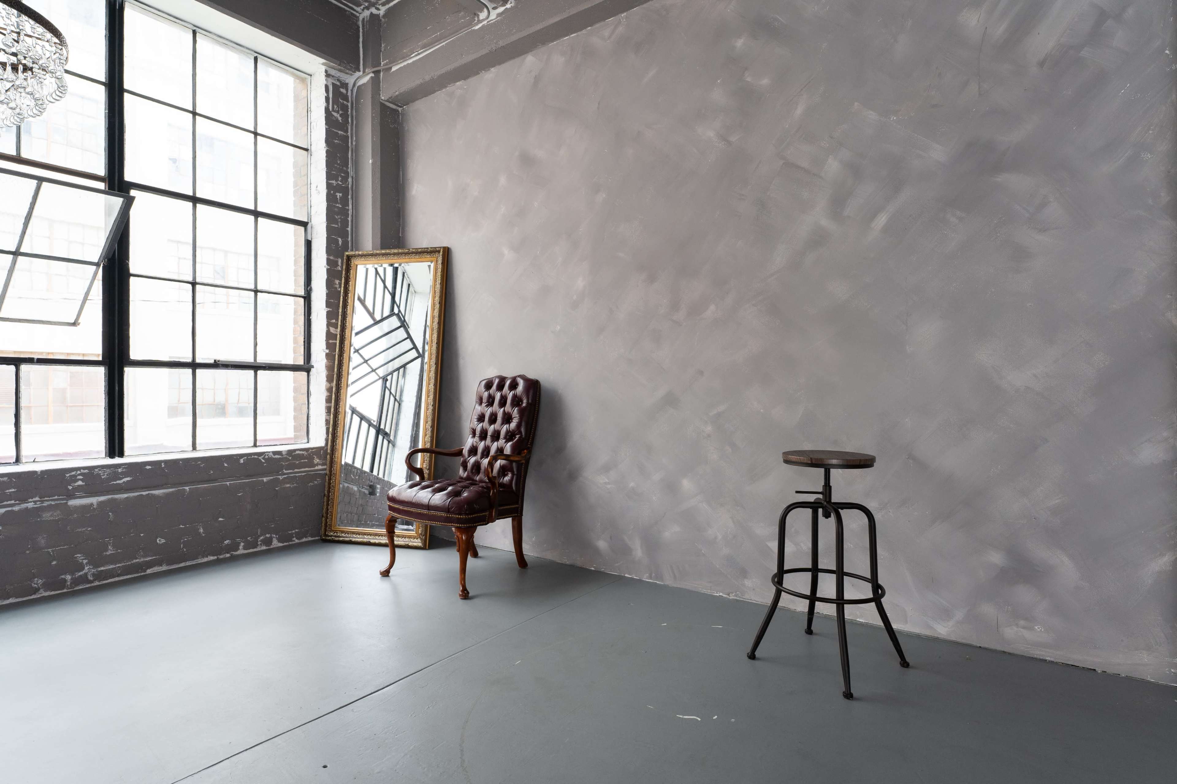 A minimalist interior with a vintage armchair and a wooden stool positioned against a gray wall, illuminated by light from large windows.