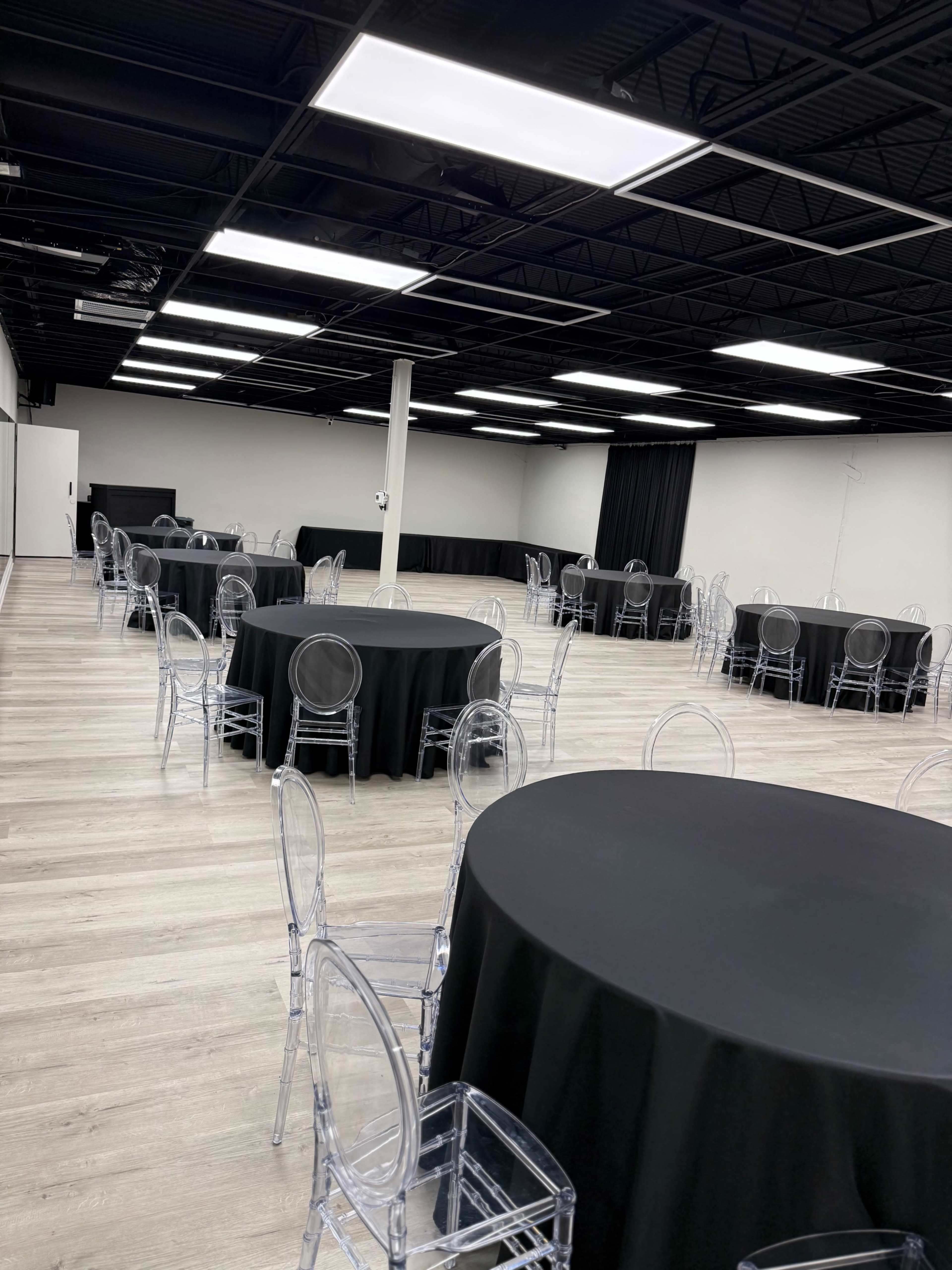 The image shows an indoor event space with several round tables covered in black tablecloths and clear acrylic chairs arranged throughout the room.
