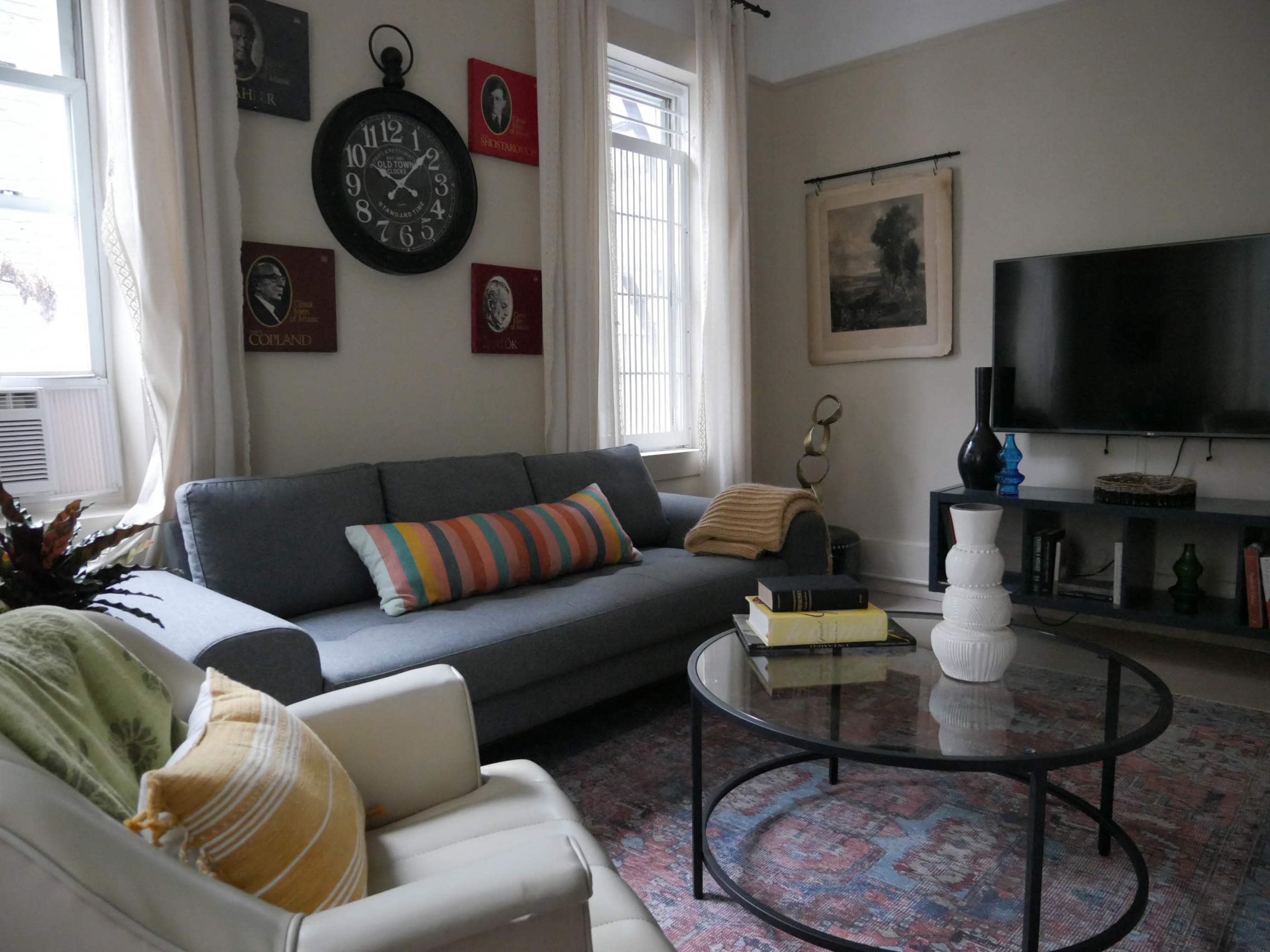 A cozy living room featuring a gray sofa with colorful cushions, a round glass coffee table, a television, and wall decor including clocks and framed art.