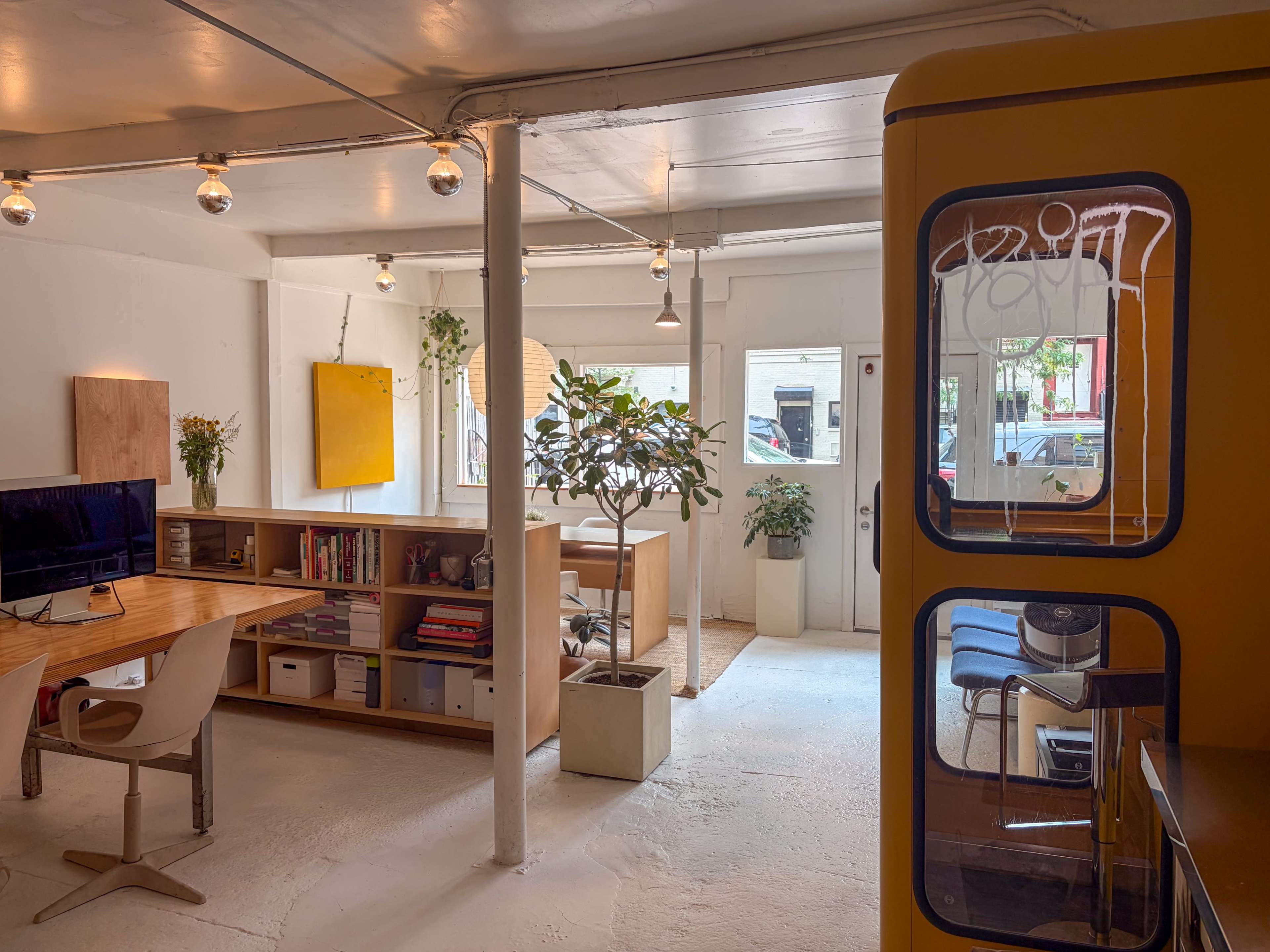 The image shows a modern, minimalist office space with wooden furniture, plants, and large windows letting in natural light.