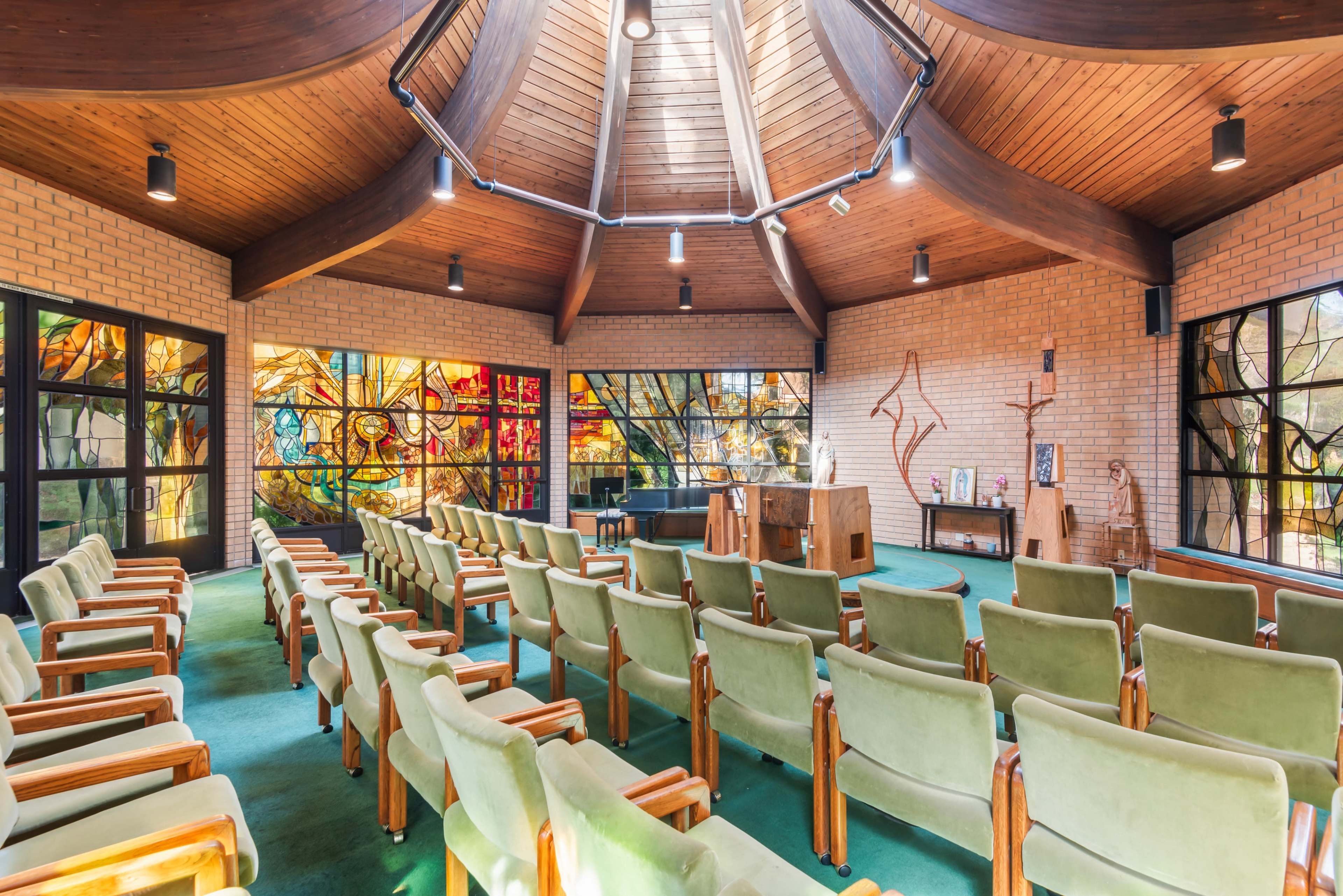 The image shows an interior of a modern chapel with wooden beams, colorful stained glass windows, and rows of green chairs facing a central altar.
