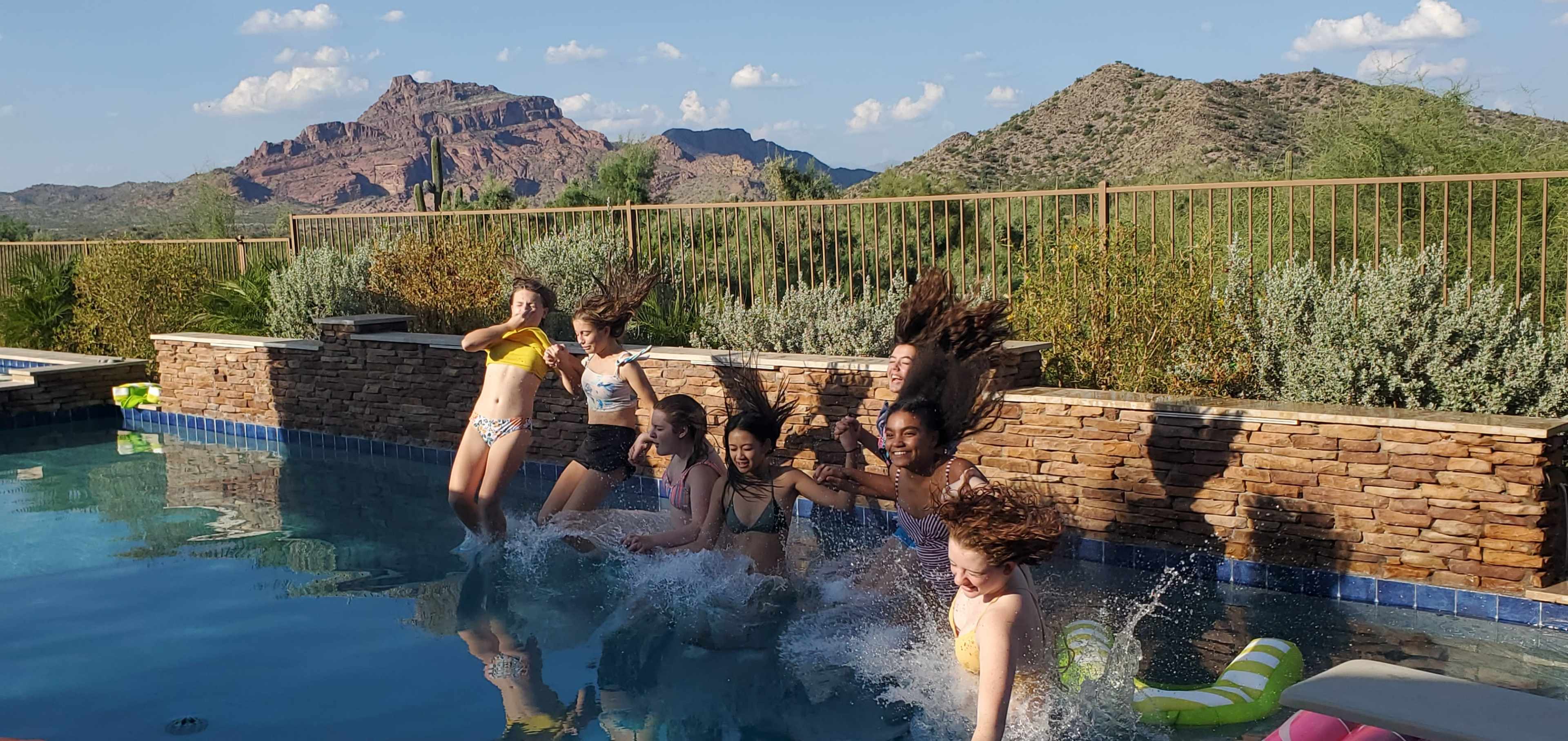 Several children jump into a swimming pool, creating splashes, with mountains in the background.