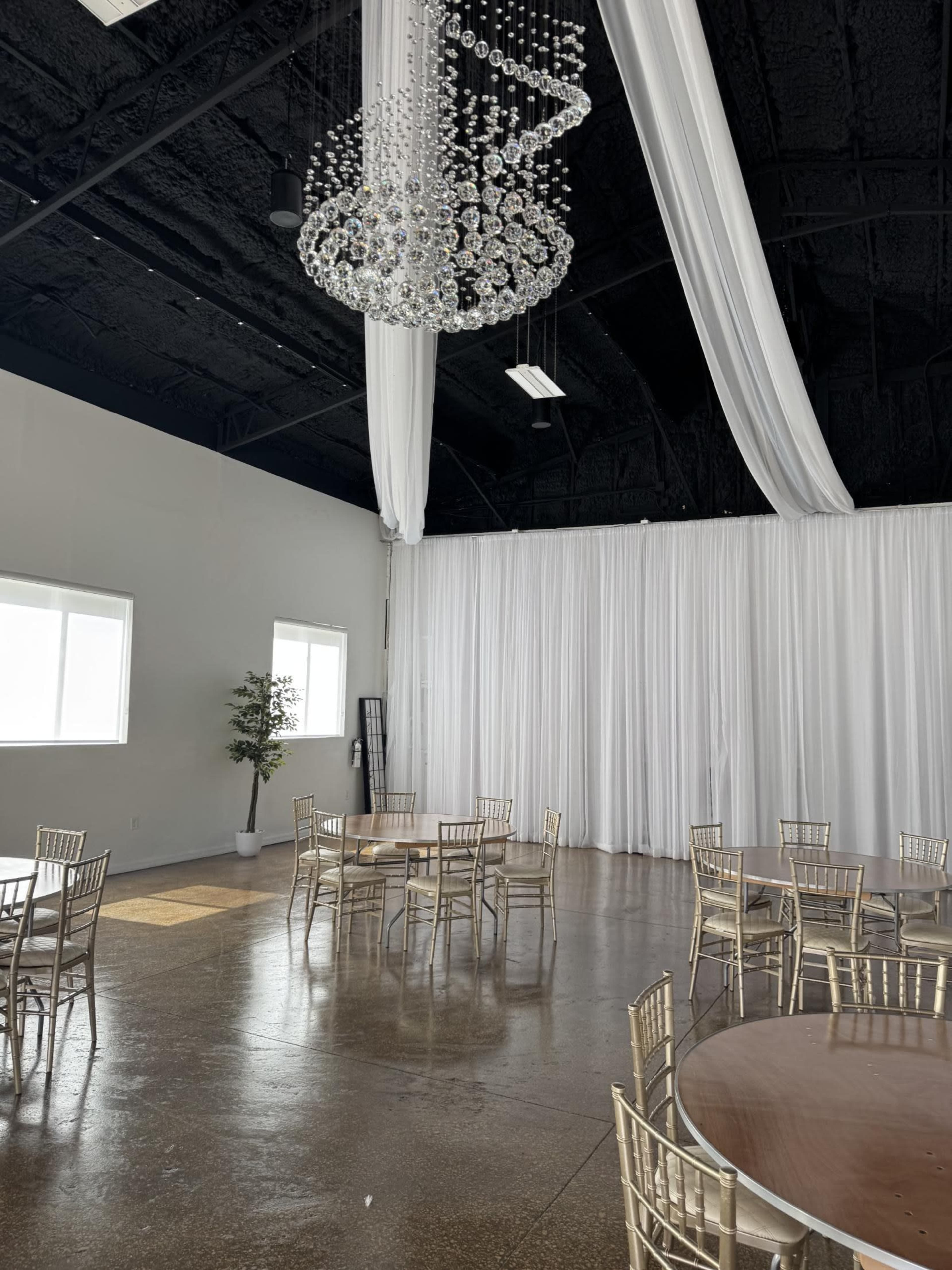 A spacious indoor venue features several round tables with gold chairs, large windows, and a decorative chandelier overhead.