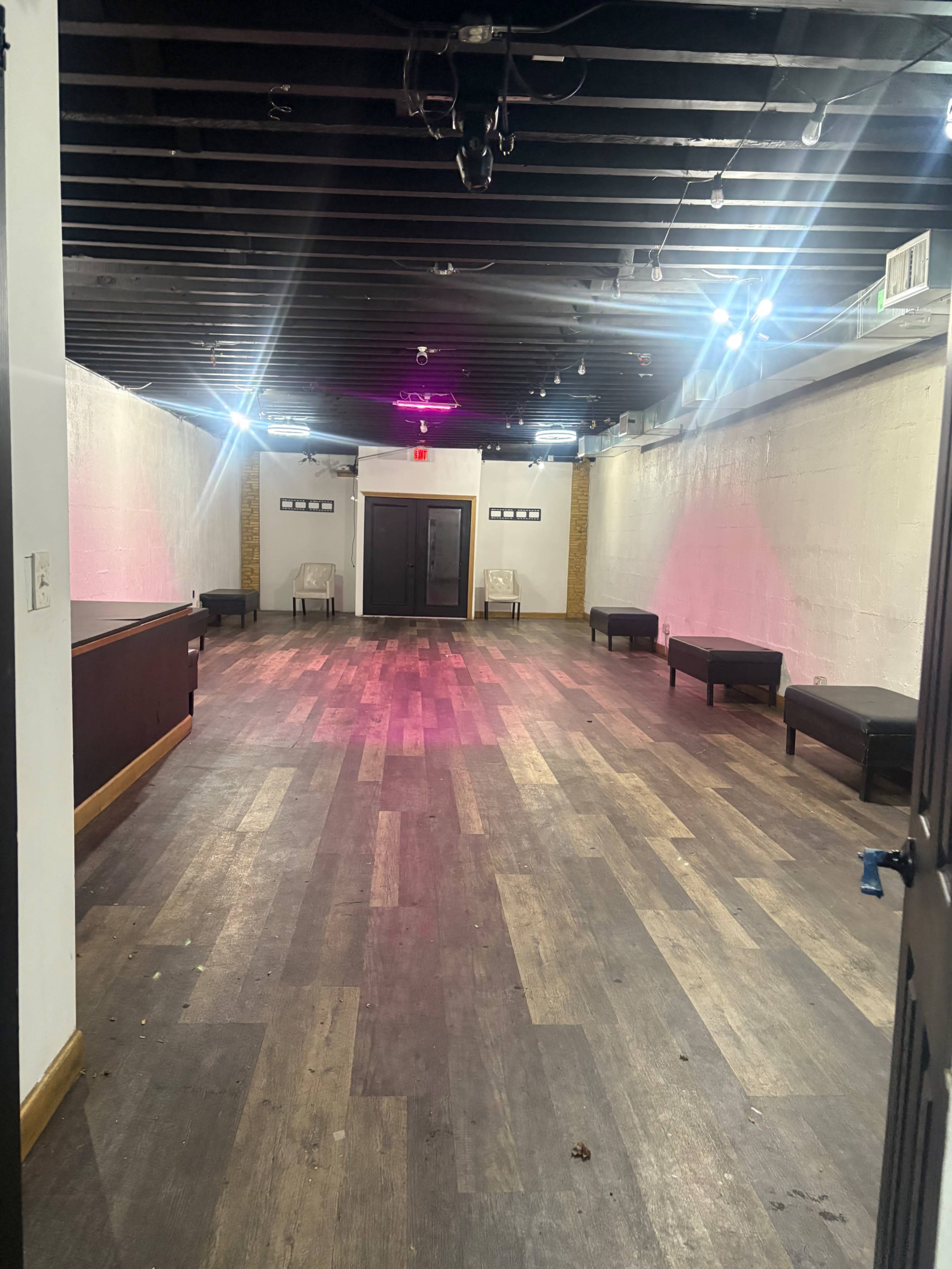 The image shows a vacant, dimly lit room with wooden flooring, two chairs against the walls, and a bar area on one side.