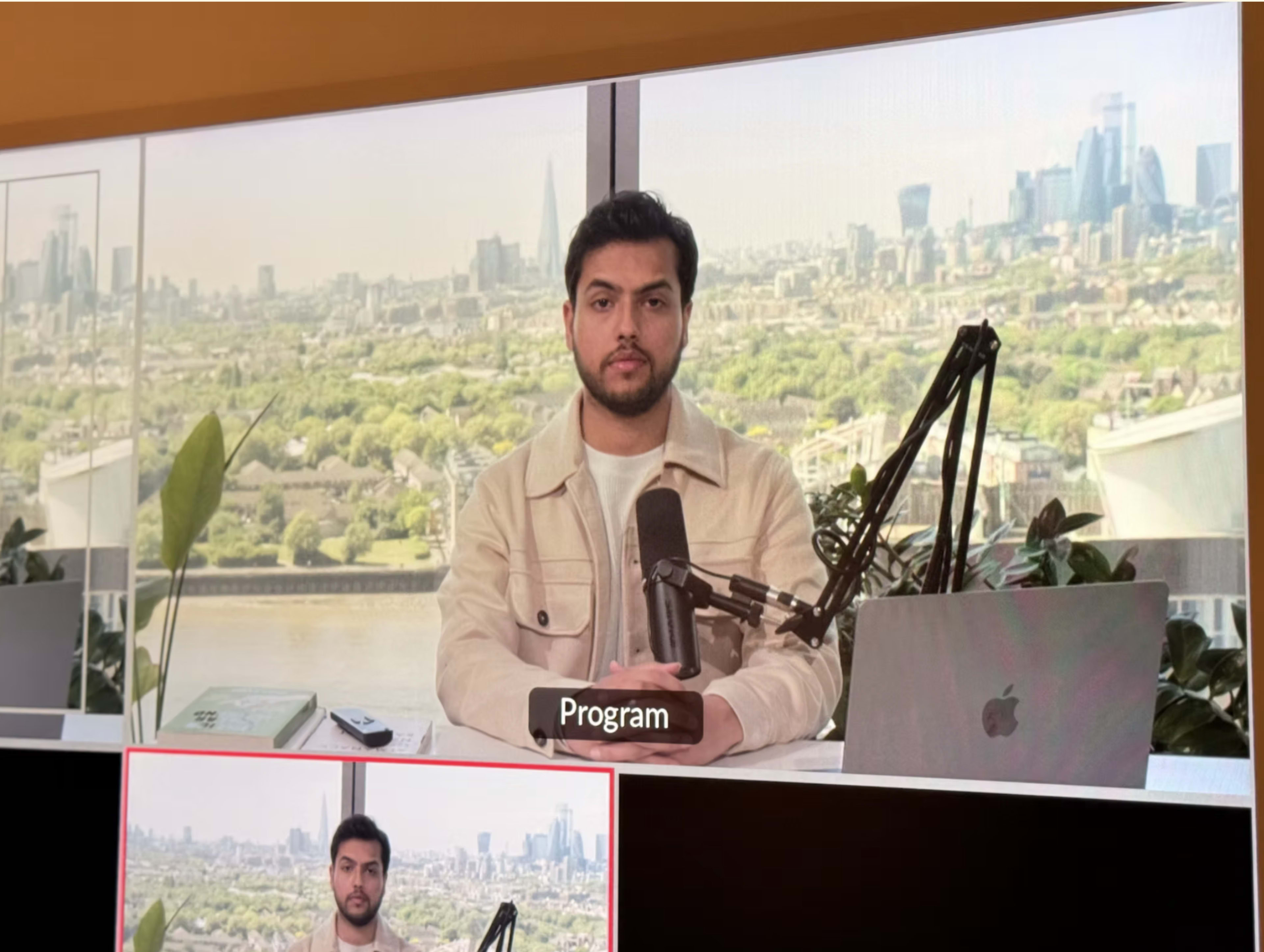 A man sits at a desk with a microphone and laptop, overlooking a city skyline through large windows.