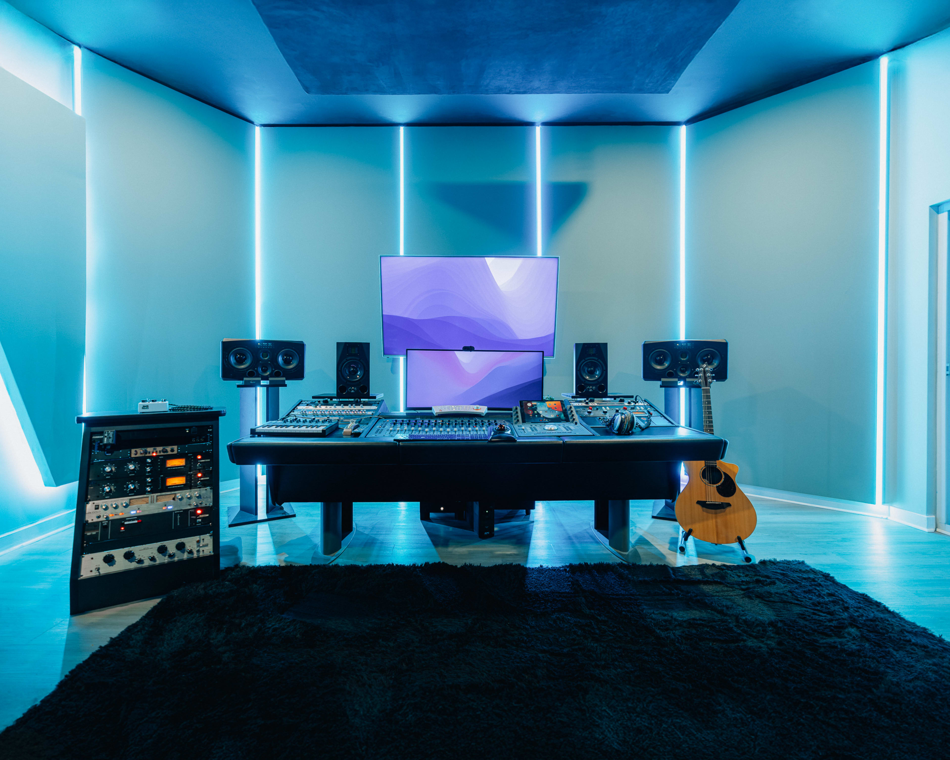 The image shows a modern music studio featuring a central desk with audio equipment, a large screen, studio monitors, and an acoustic guitar against a backdrop of illuminated wall panels.