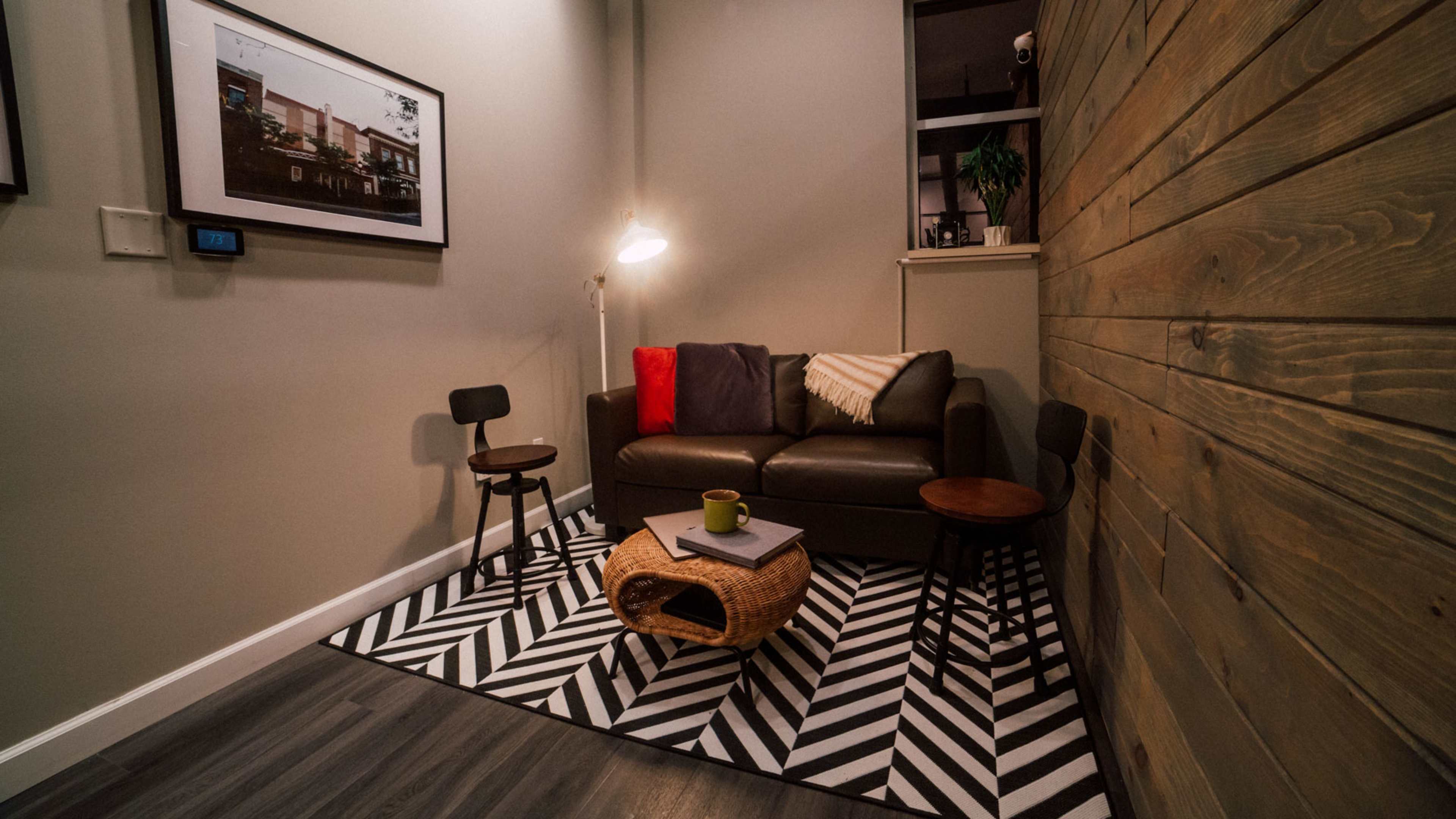 A small sitting area with a brown sofa, two wooden chairs, a striped rug, and a coffee table, all set against a textured wooden wall.